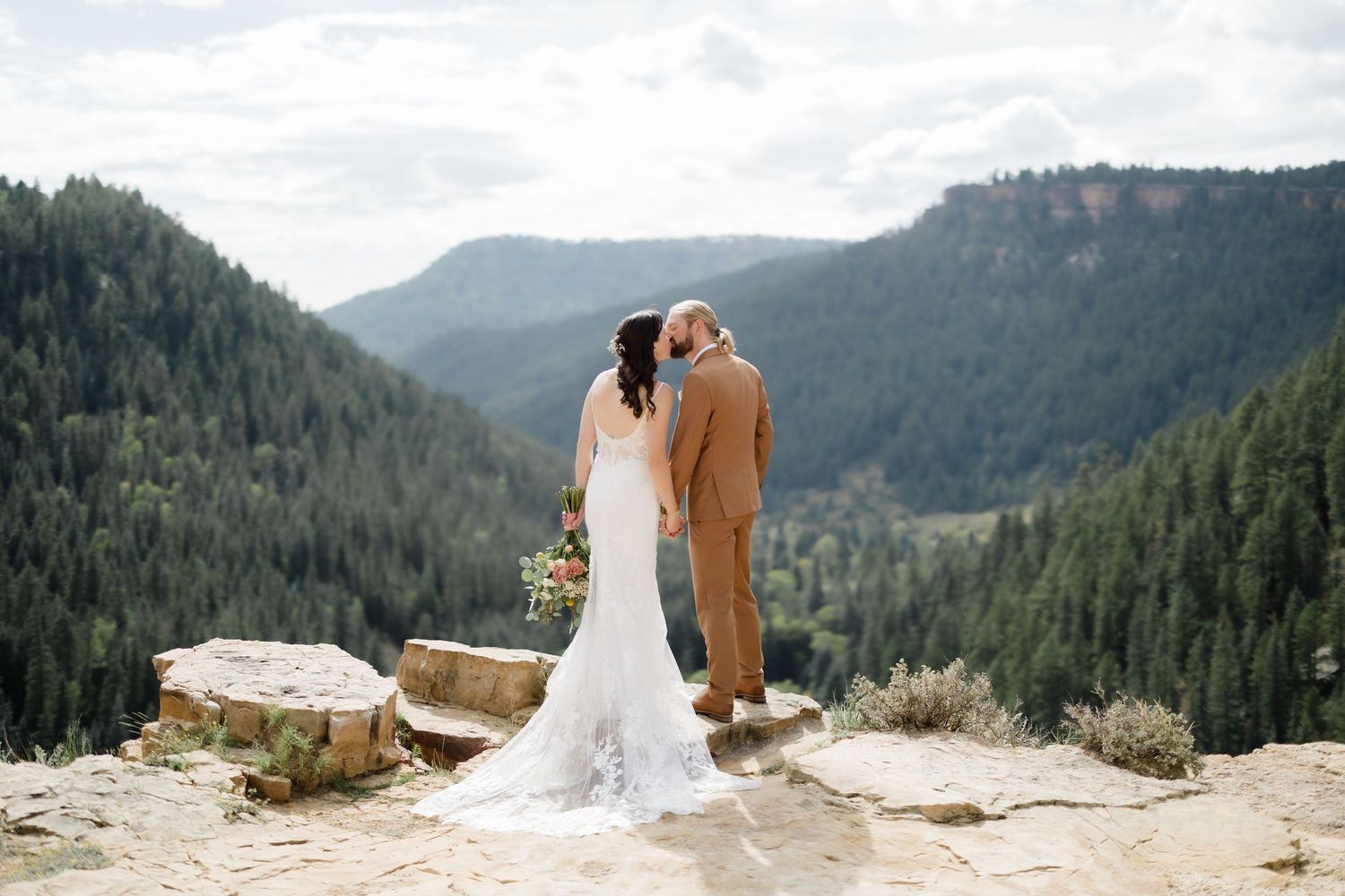 Bride and groom share a kiss overlooking a stunning mountain valley with lush pine forests on their wedding day.