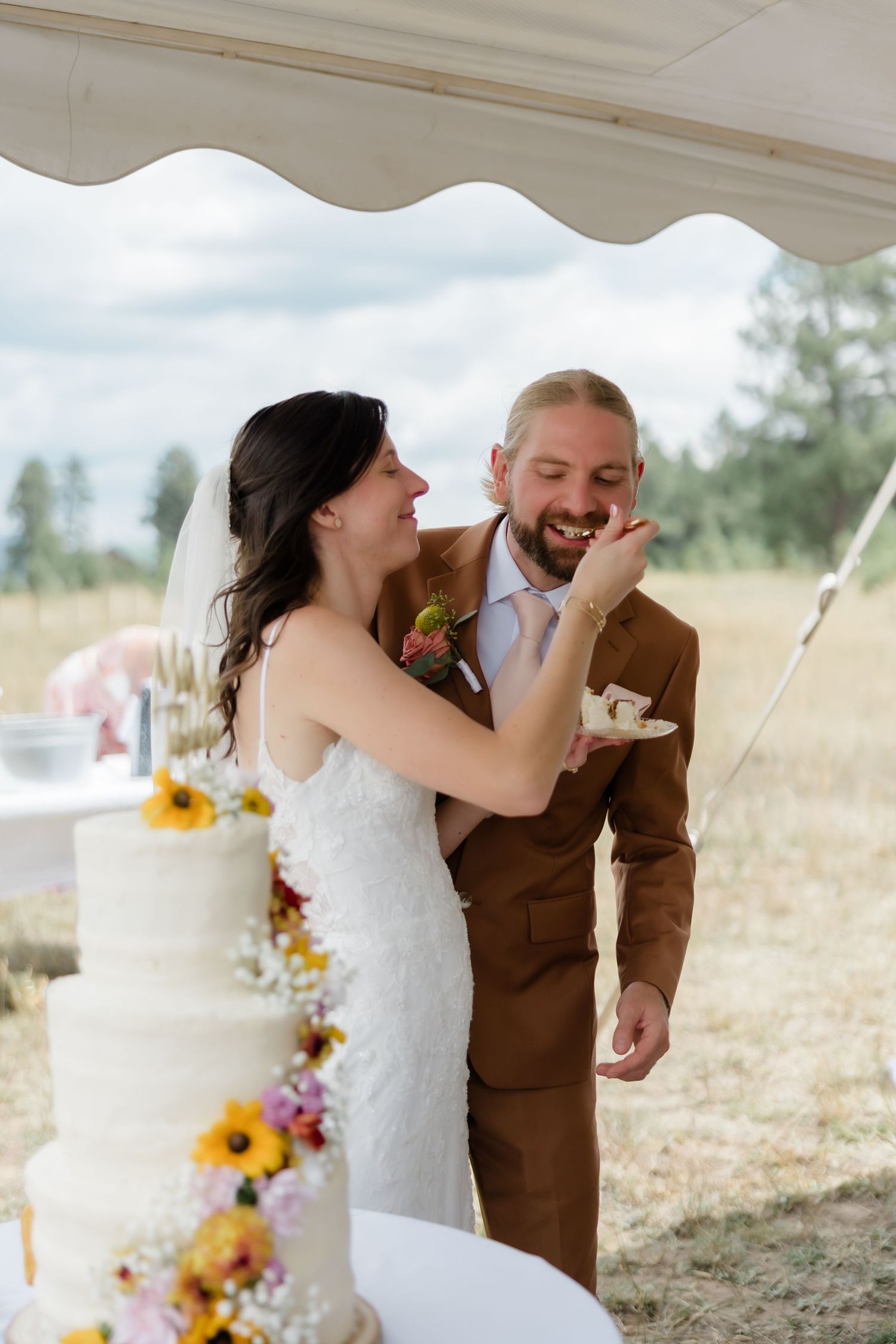 Bride feeding wedding cake to groom in brown suit outdoors, with a white floral tiered cake in the foreground.
