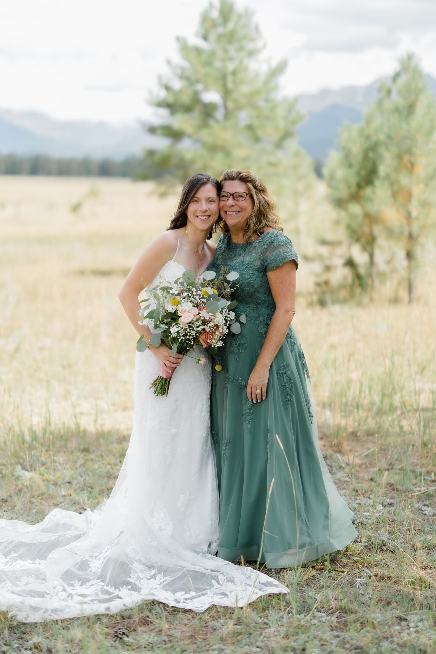 Bride in white gown and woman in green dress pose outdoors with bouquet, mountains and pine trees behind them.