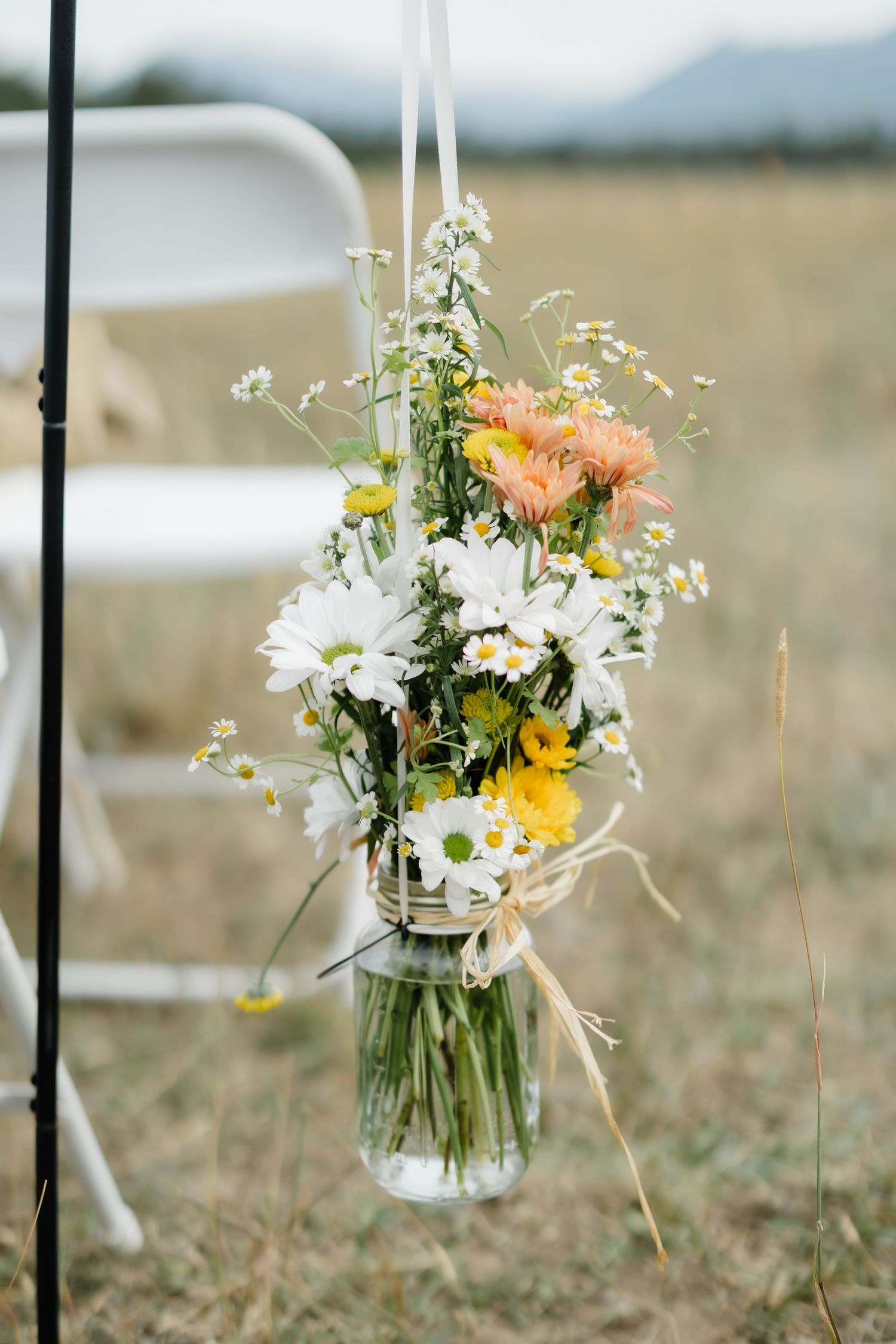Wildflower bouquet in mason jar hanging outdoors at rustic wedding ceremony with daisies and gerberas.