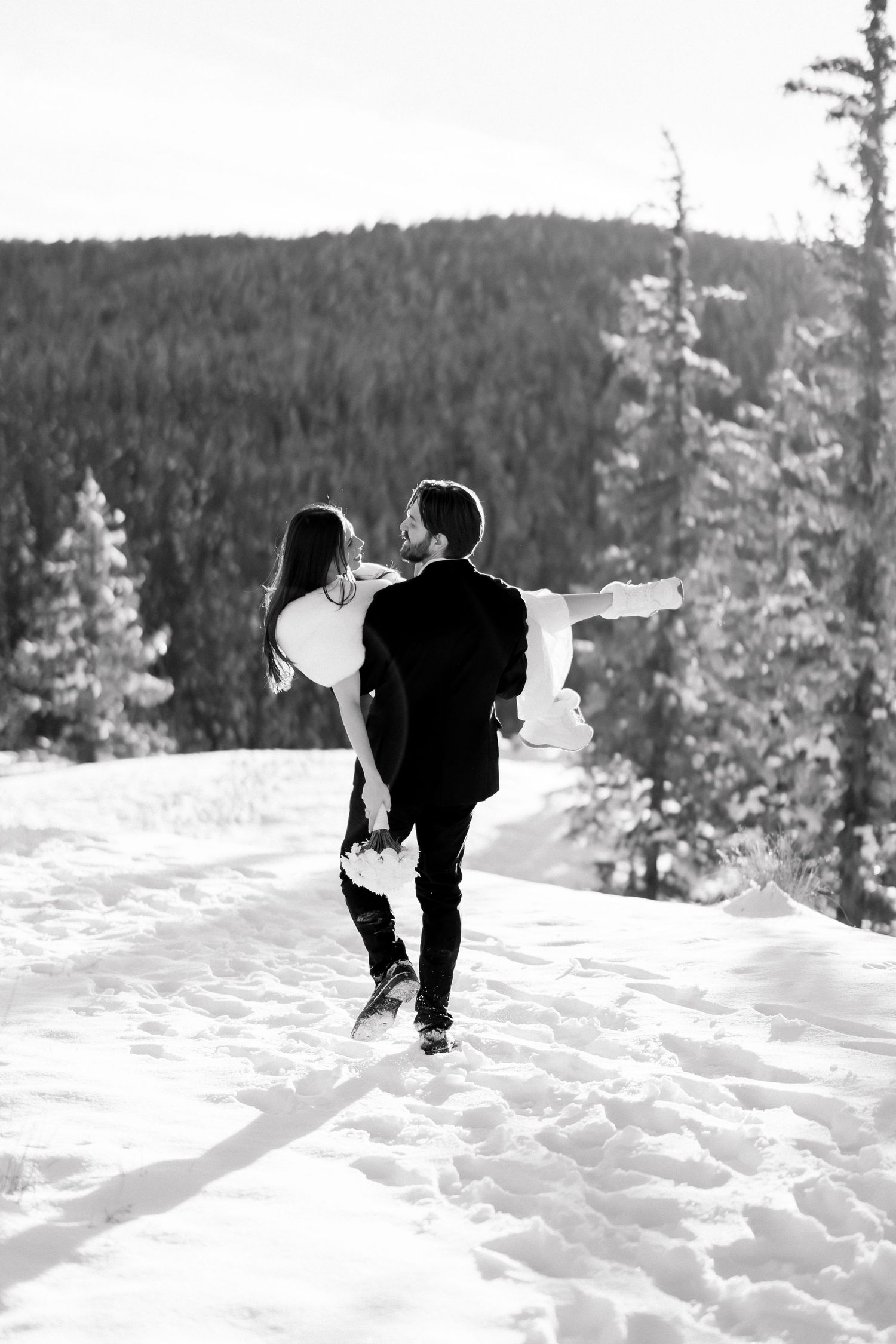 Groom carries bride through snowy mountain landscape with pine forest backdrop, black and white photo.