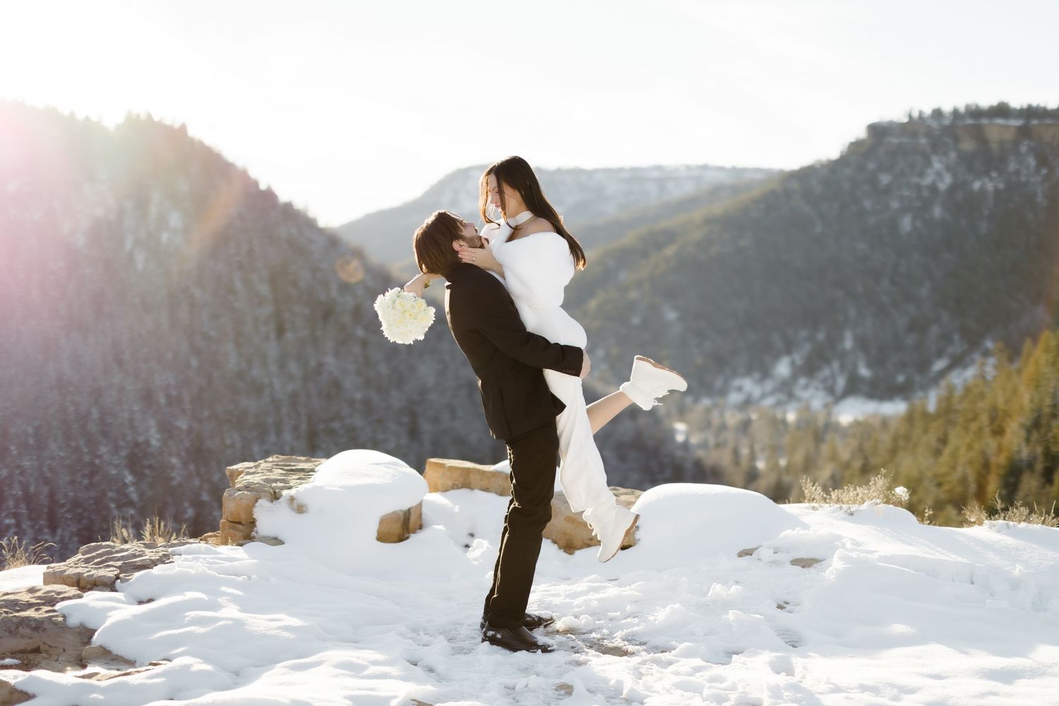 Groom lifting bride in snowy mountain landscape during winter wedding photo session with sunlit peaks behind.