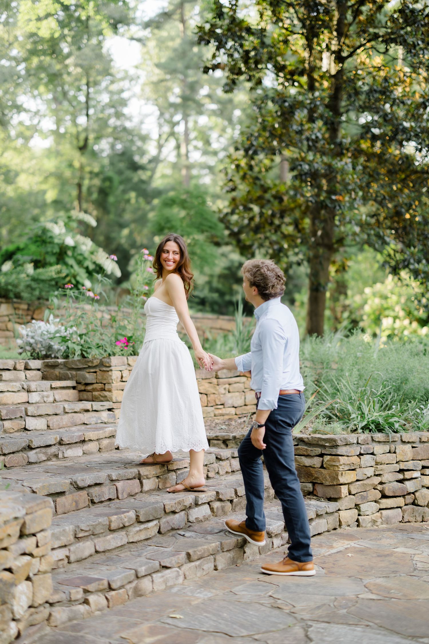 Couple holding hands on stone steps in a lush garden, woman in white dress smiling back at man in light blue shirt.
