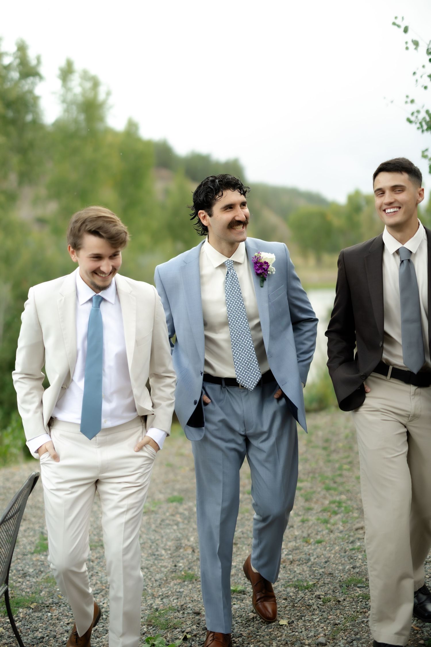 Three groomsmen in suits and ties walk outdoors, smiling, with the groom in a light blue suit wearing a boutonniere.