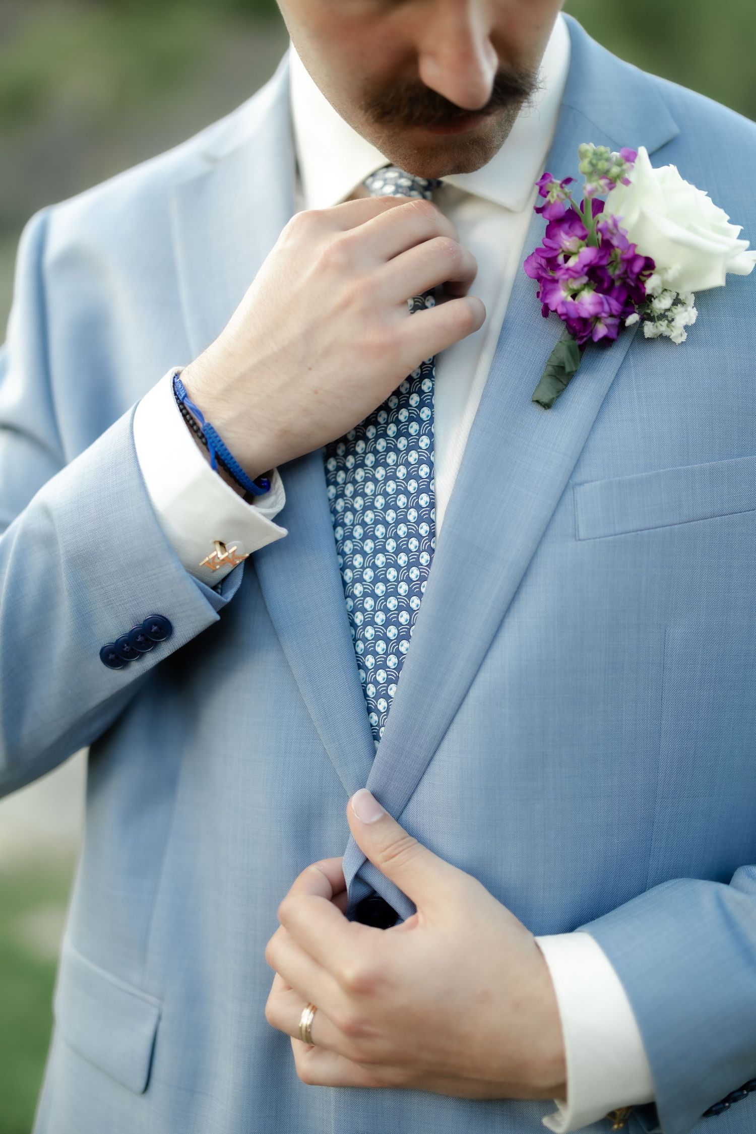 Close-up of groom in blue suit straightening tie, wearing cufflinks and wedding ring with floral boutonniere.