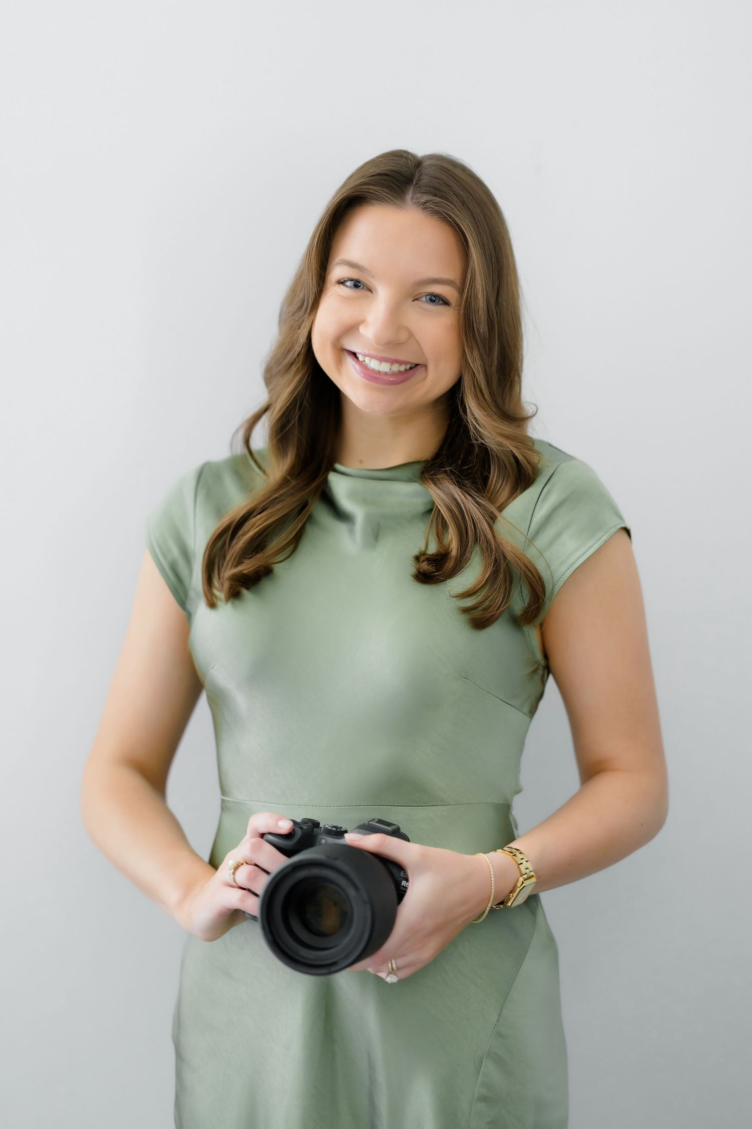 Smiling woman in sage green dress holding a professional camera, portrait photographer branding photo on white background.