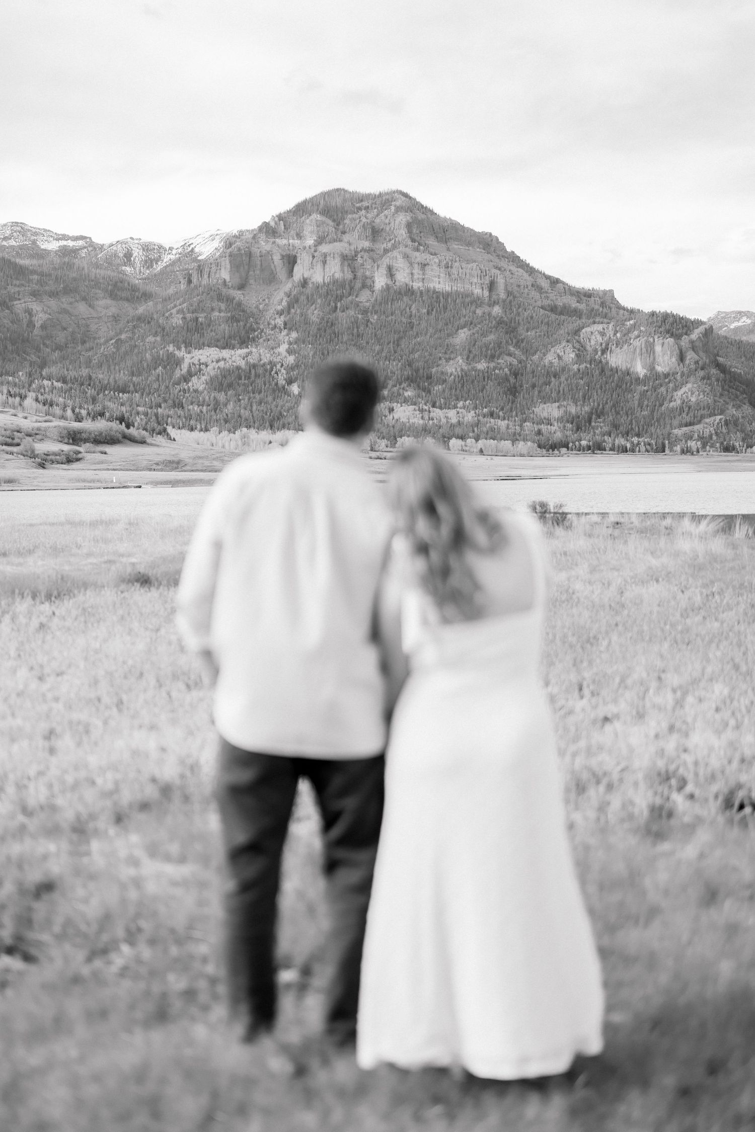 Black and white photo of couple holding hands gazing at mountain landscape and lake in field.