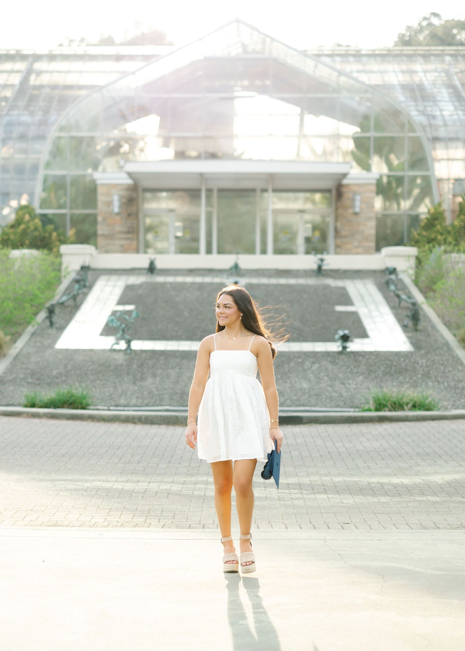High school senior in white dress walking near ornate greenhouse building at Birmingham Botanical Gardens in Birmingham, Alabama.