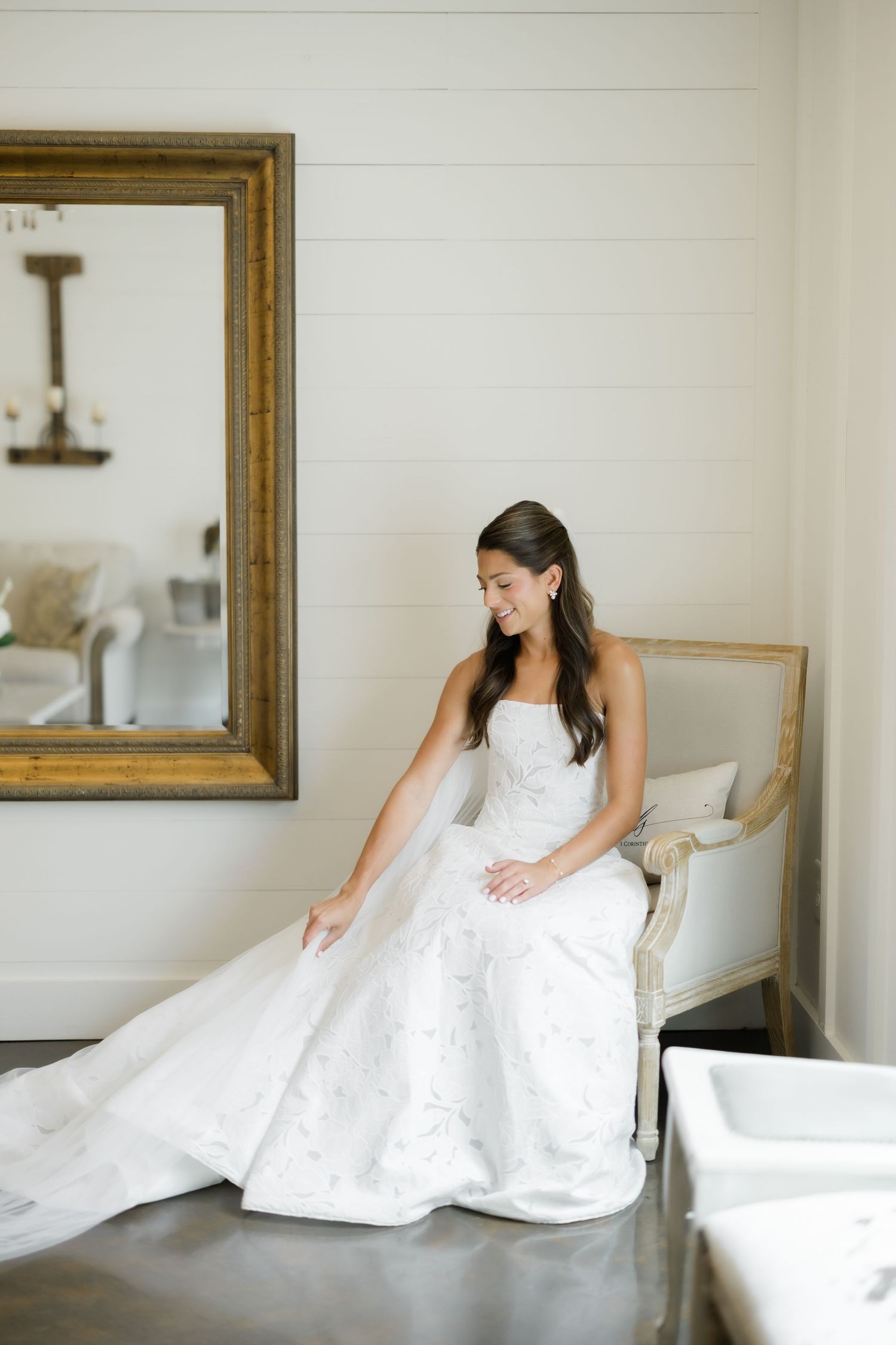 Bride in strapless white wedding gown with floral detail sits on vintage chair beside large gold mirror.