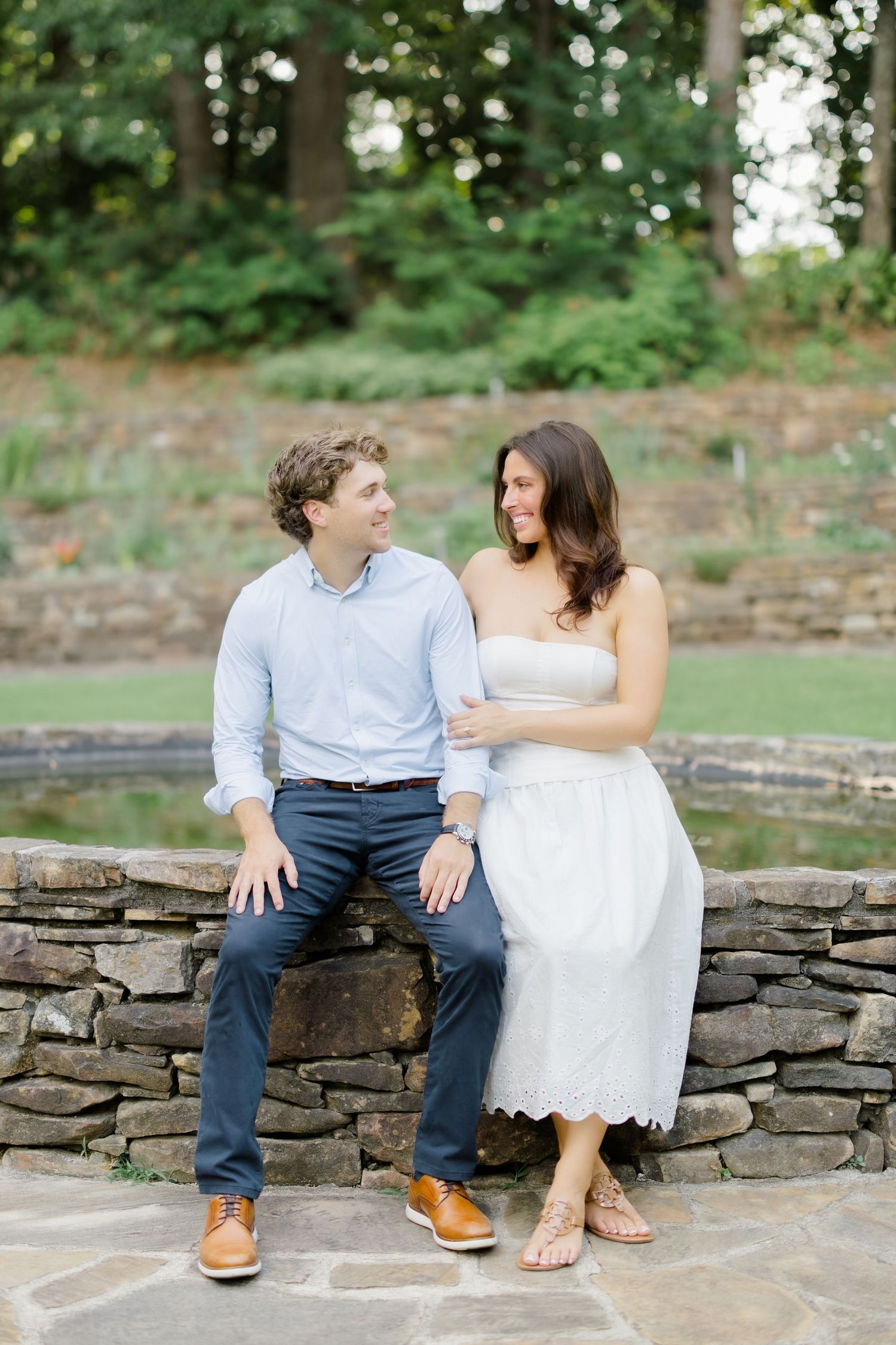 Couple sitting on a rustic stone wall smiling at each other, man in blue shirt, woman in white strapless dress.
