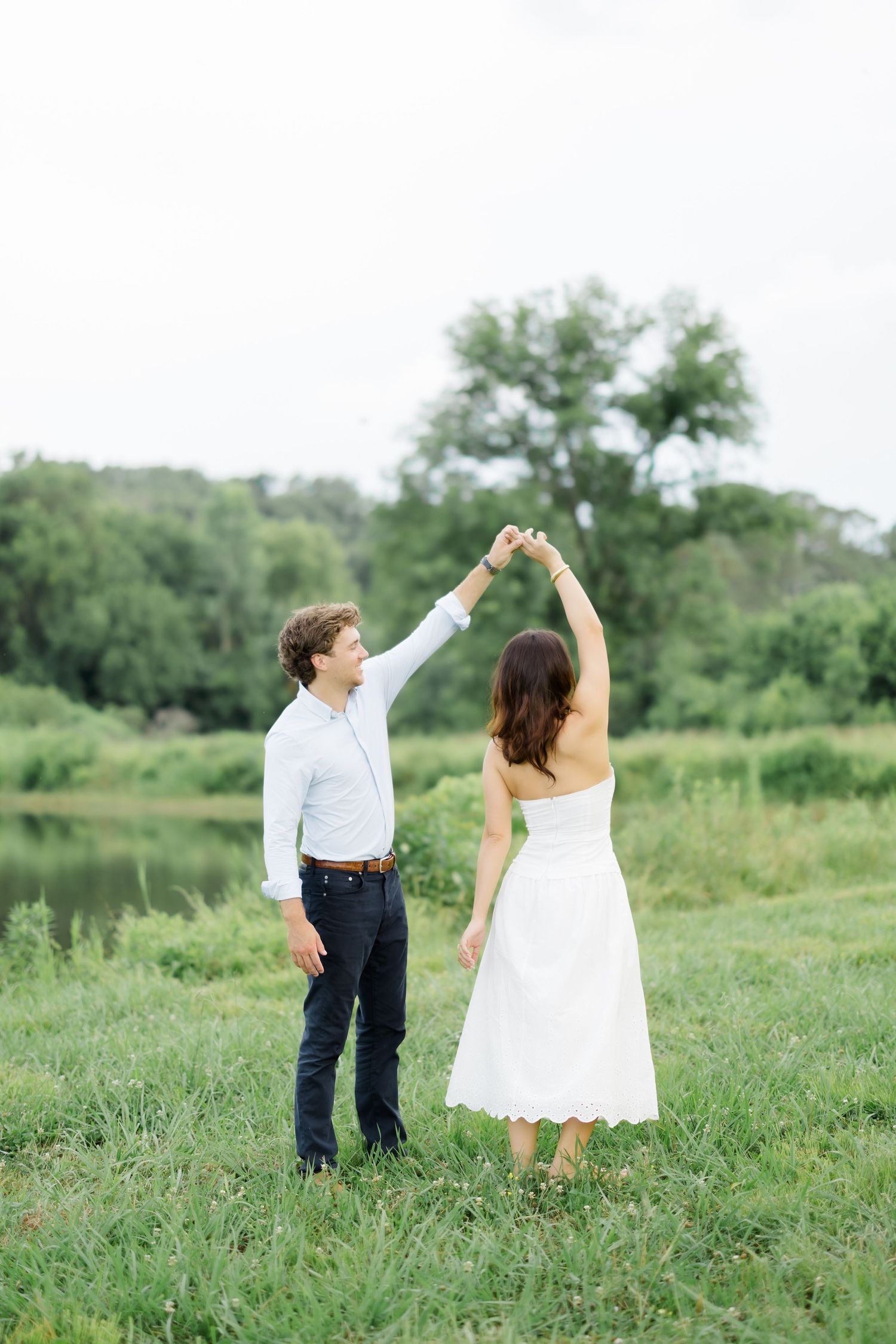 A couple dances together in a lush green field near a pond, the man spinning the woman in a white dress.