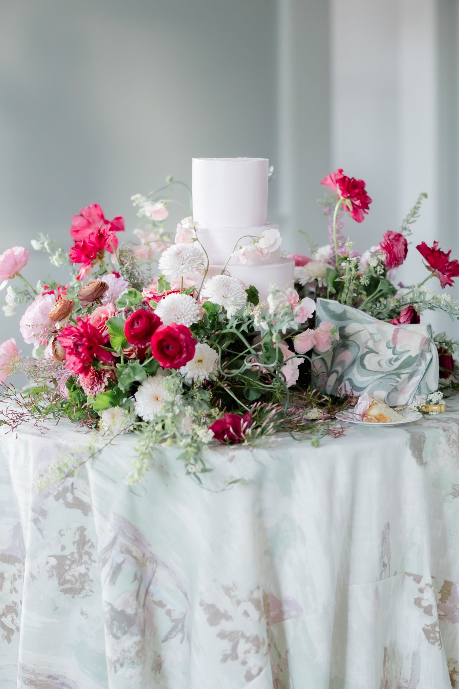 Wedding cake table decorated with pink and red flowers, white linens, and greenery for an elegant wedding reception.