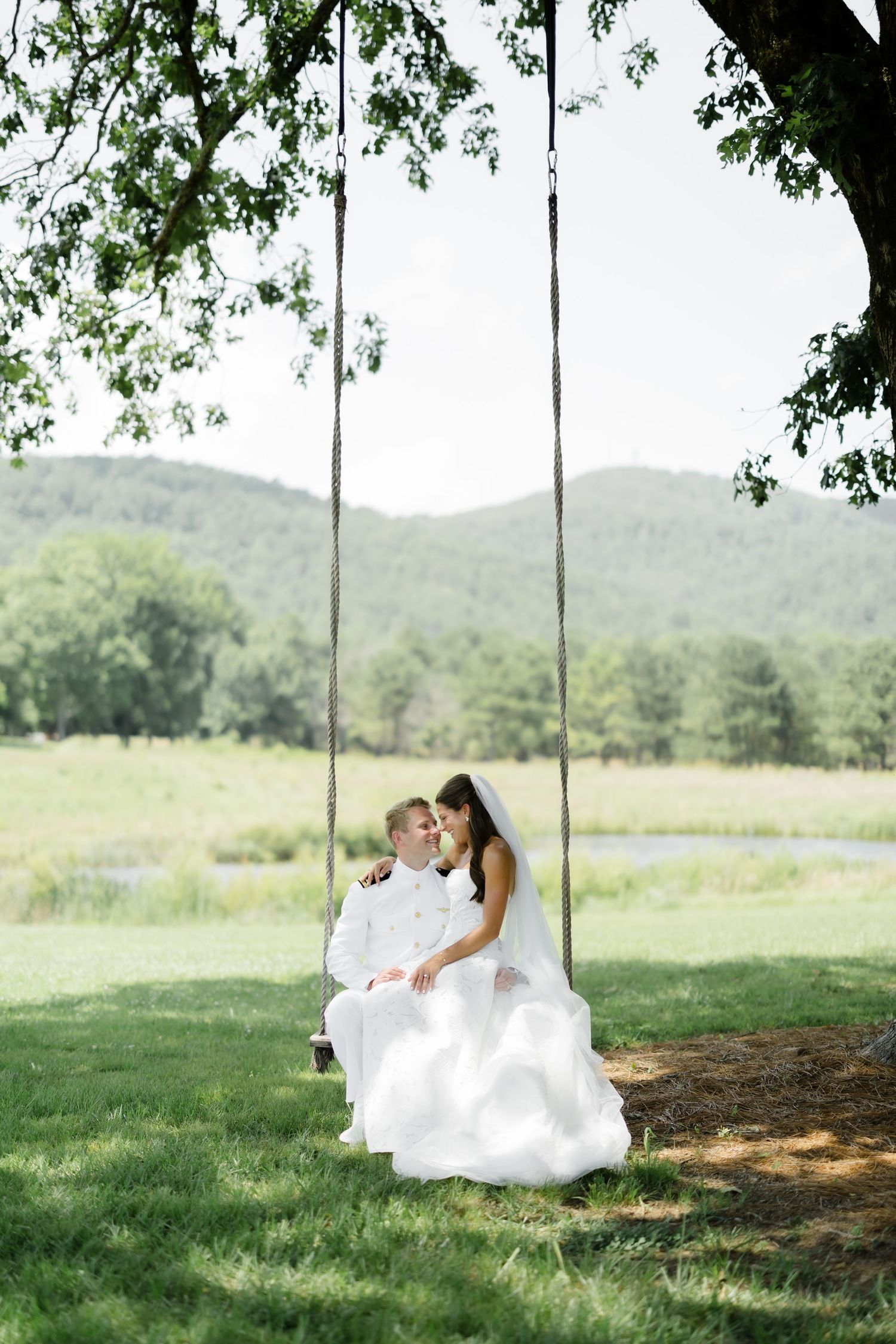 Bride and groom sitting together on a rope swing under a large tree with scenic green hills in the background.