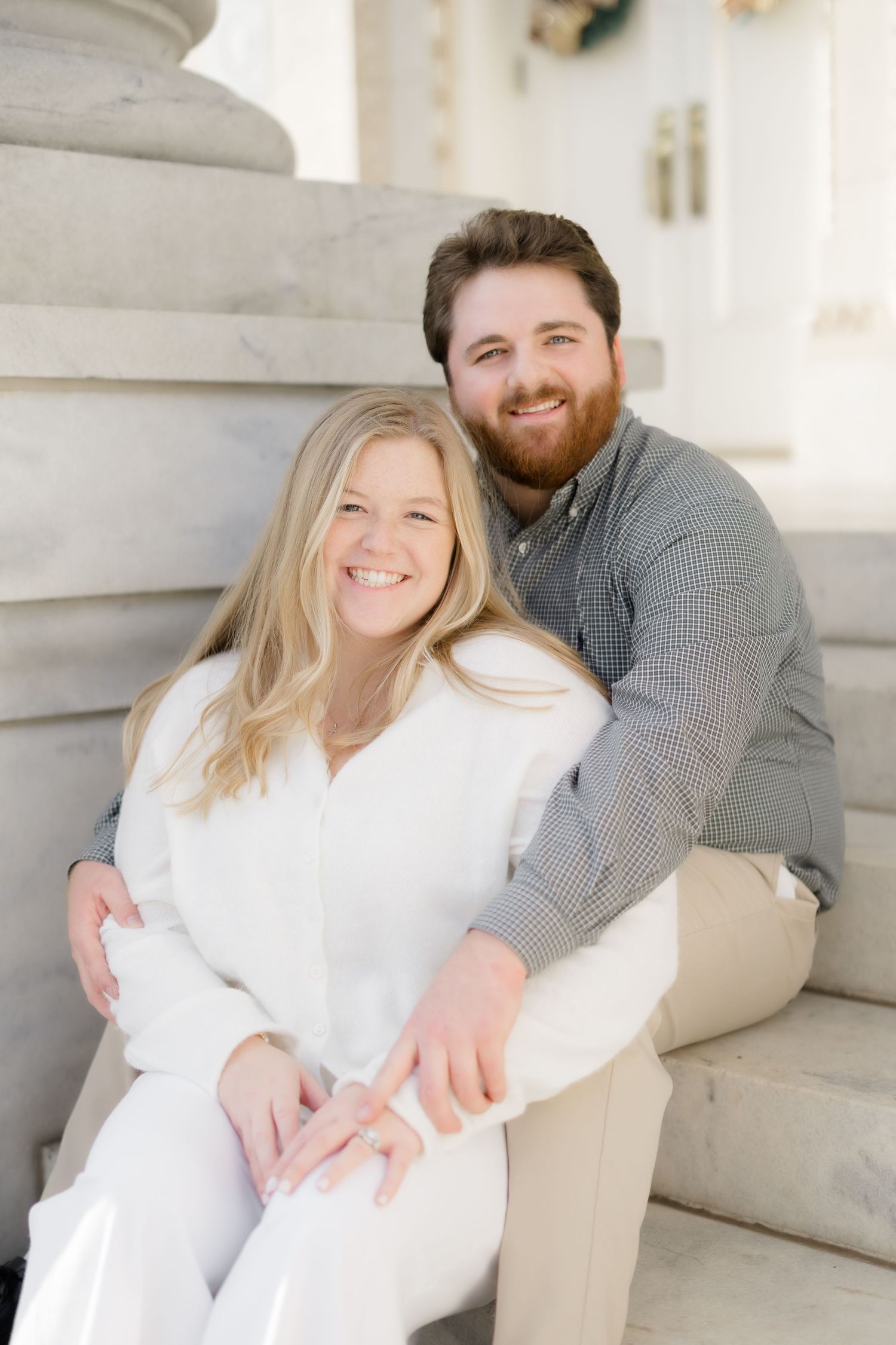 A smiling couple sits on stone steps, the woman in white and the man in a gray shirt, posing for an engagement photo.