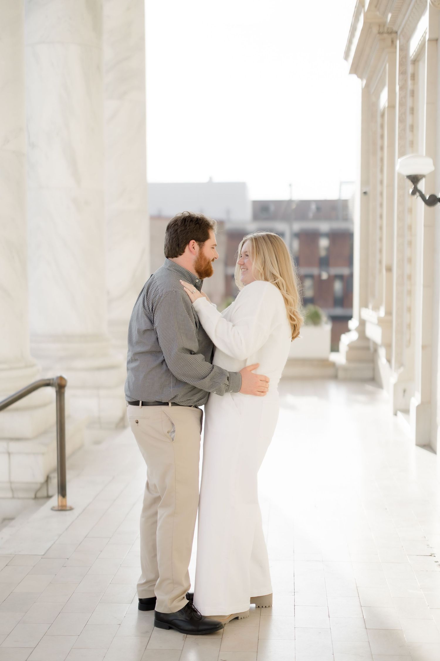 A couple stands close together, gazing lovingly at each other in a sunlit marble colonnade during an engagement session.