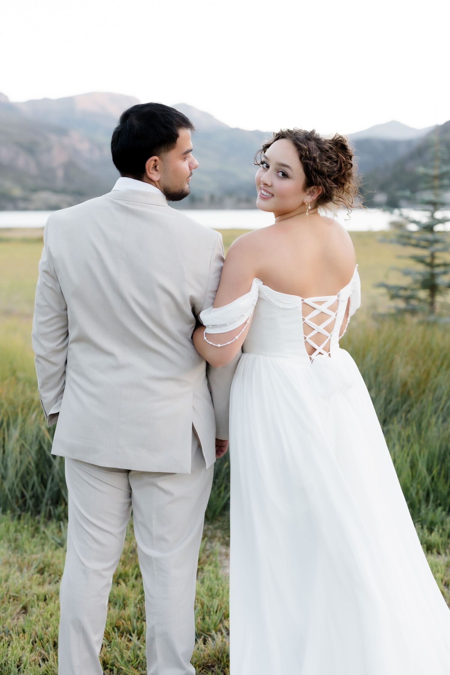 Bride in white off-shoulder gown with lace-up back stands with groom in beige suit, mountain lake backdrop.