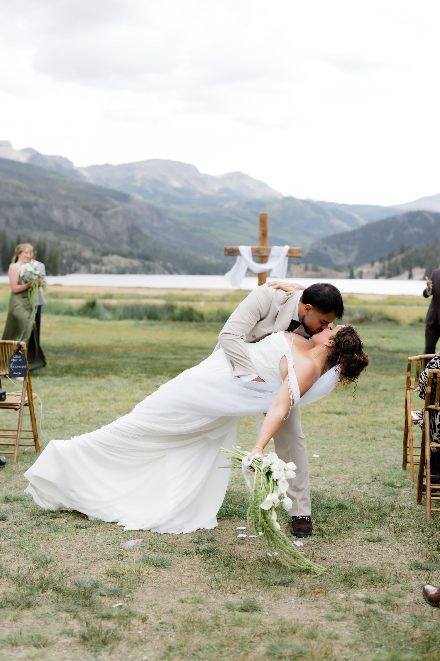 Groom dipping bride for a romantic kiss after outdoor mountain wedding ceremony with wooden cross backdrop.