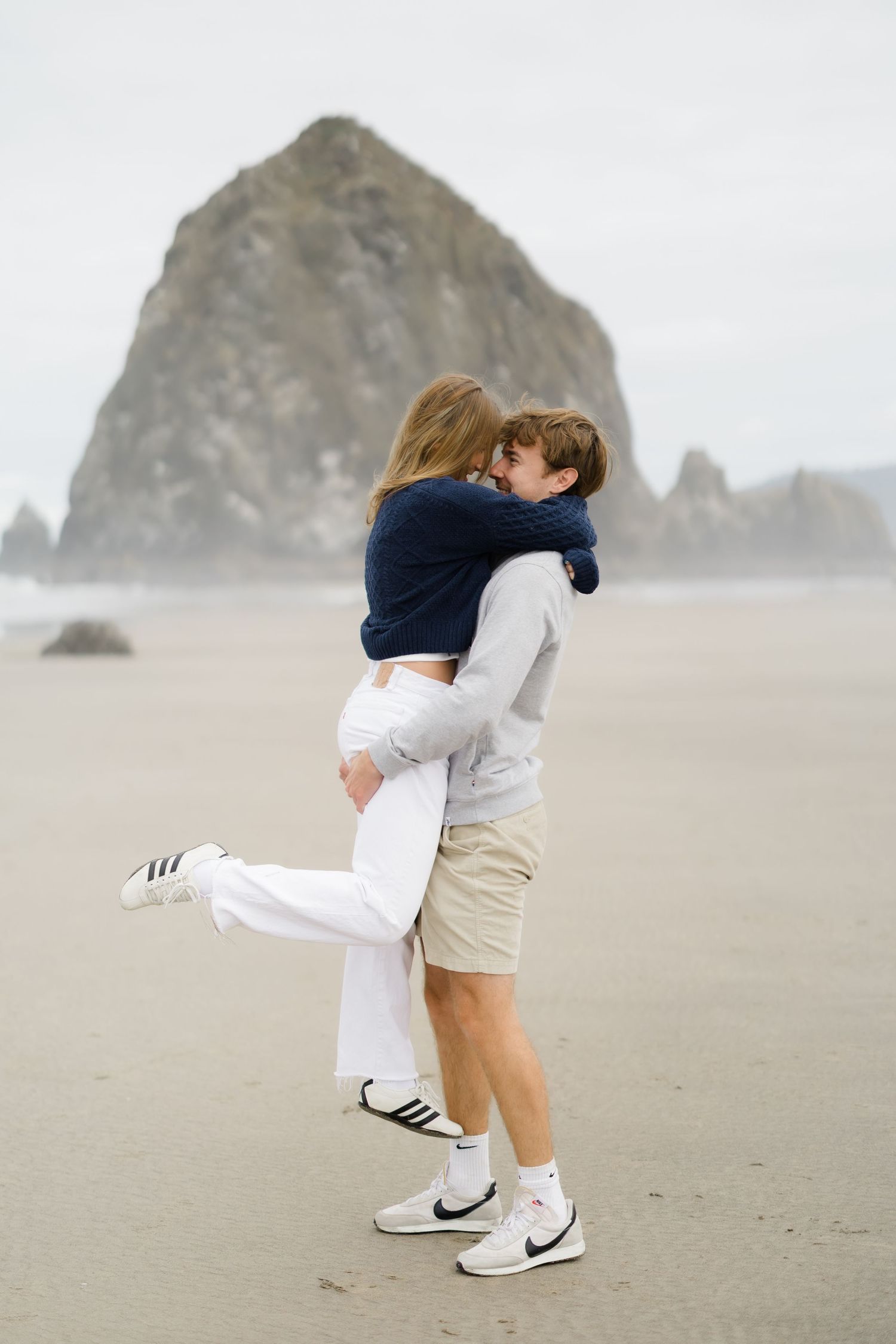Couple sharing a romantic kiss on a misty beach with Haystack Rock in the background at Cannon Beach, Oregon.