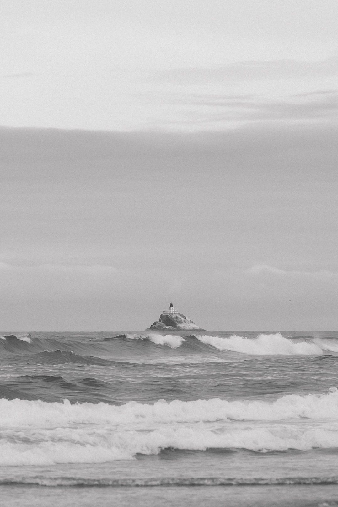 Black and white ocean seascape with crashing waves and a small rocky island with a lighthouse in the distance.