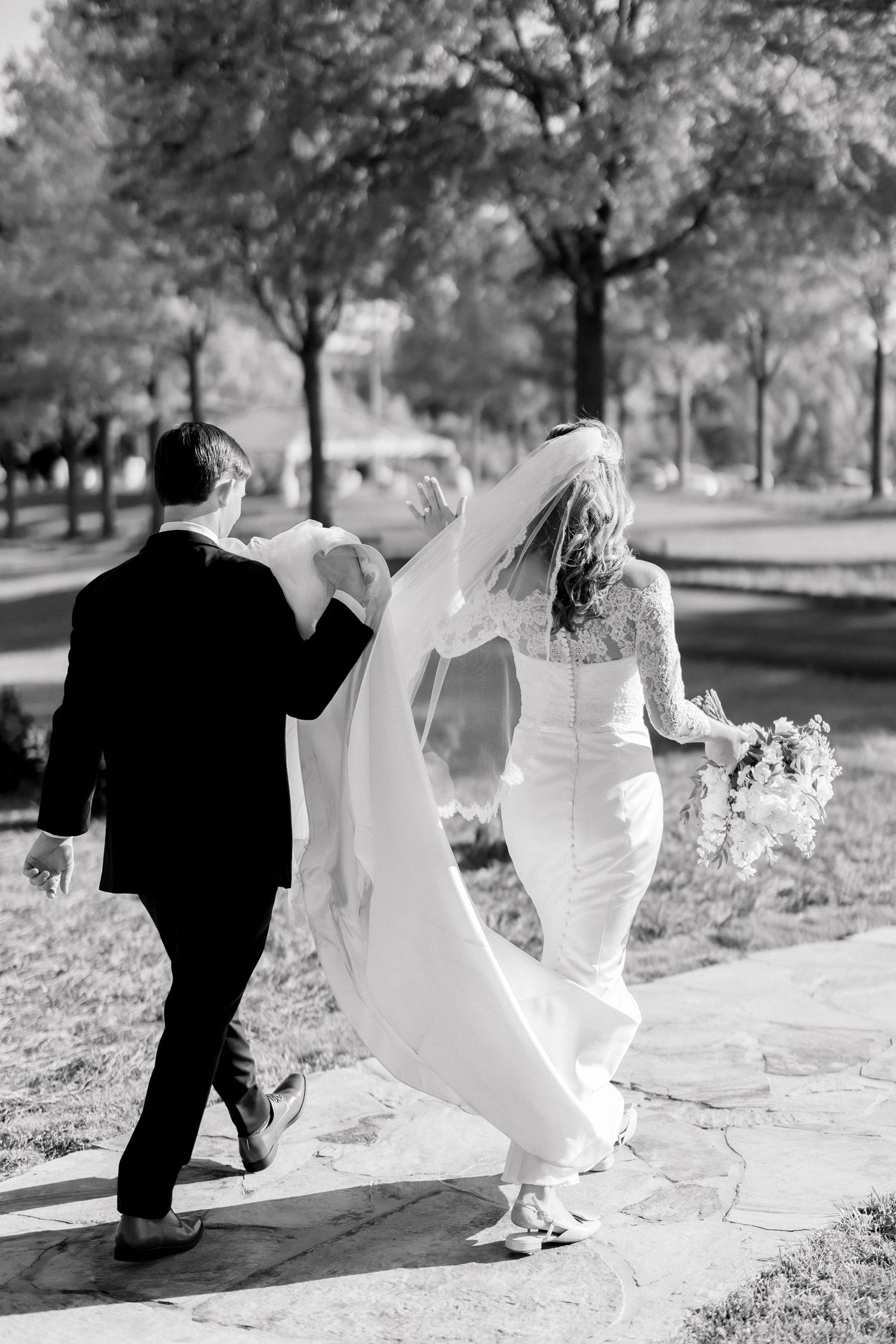 Black and white photo of bride and groom walking hand in hand outdoors, bride holding bouquet in flowing wedding gown.
