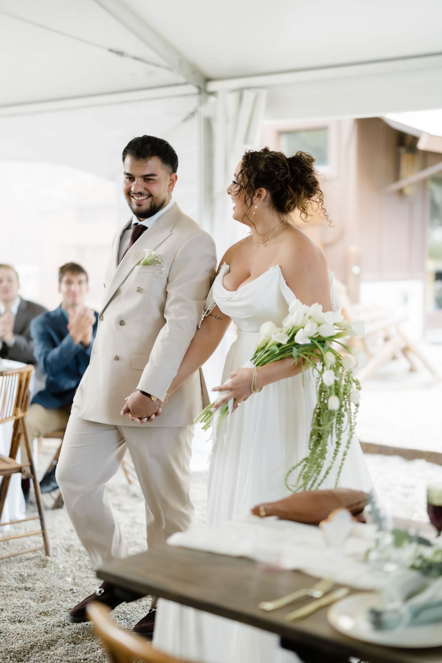 Smiling groom in beige suit holds brides hand as they walk together at their wedding ceremony.