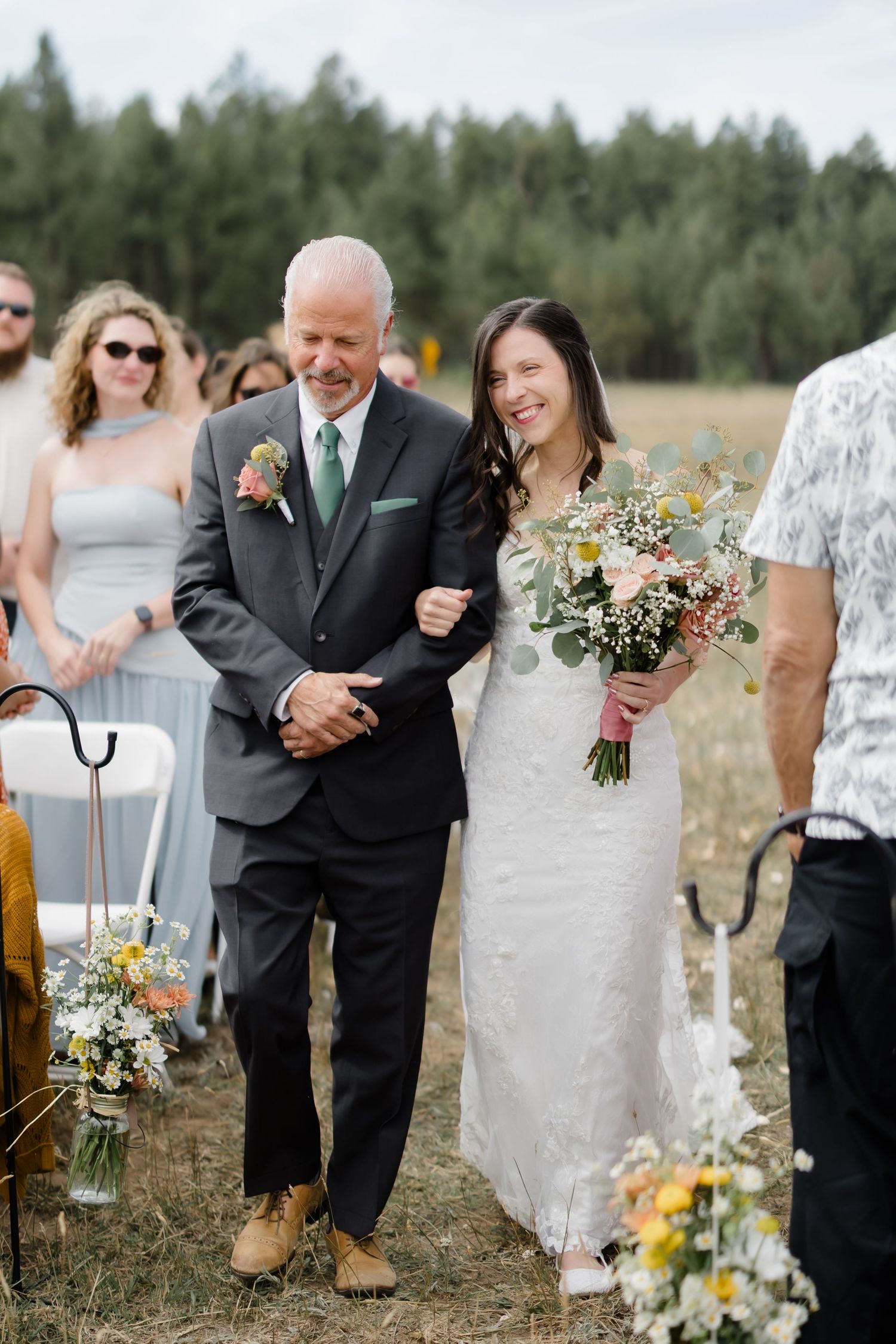 Bride in white gown holding bouquet walks down outdoor aisle with older man in gray suit at rustic wedding.