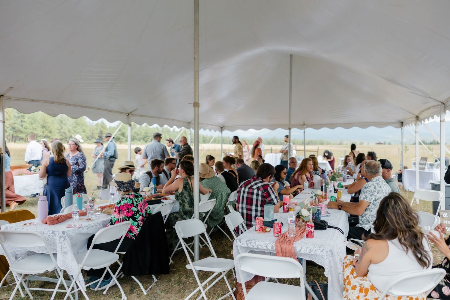 Guests gather under a large white tent for an outdoor event, seated at round tables with white chairs.