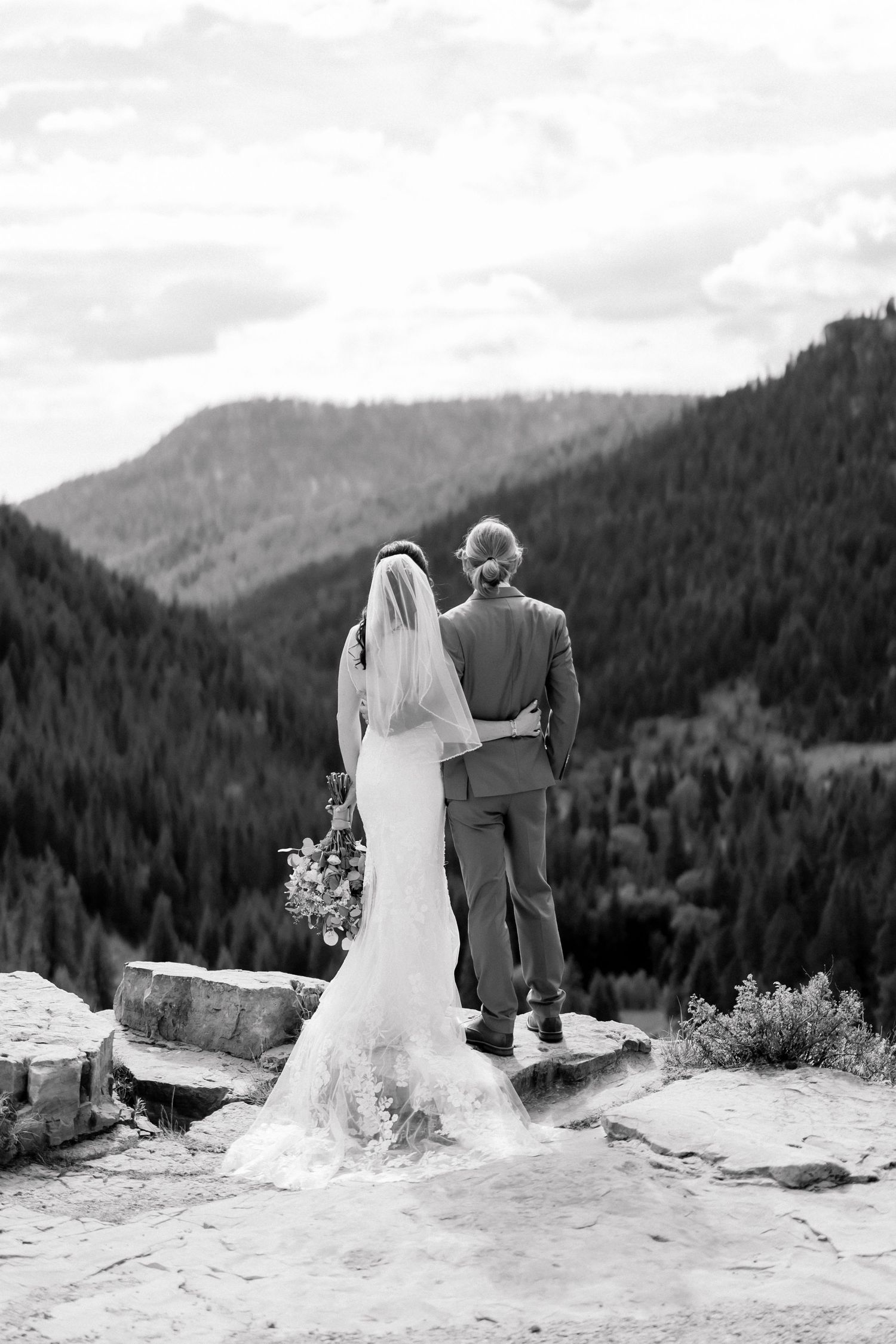 Black and white photo of bride and groom standing on rocks overlooking stunning mountain forest landscape on wedding day.