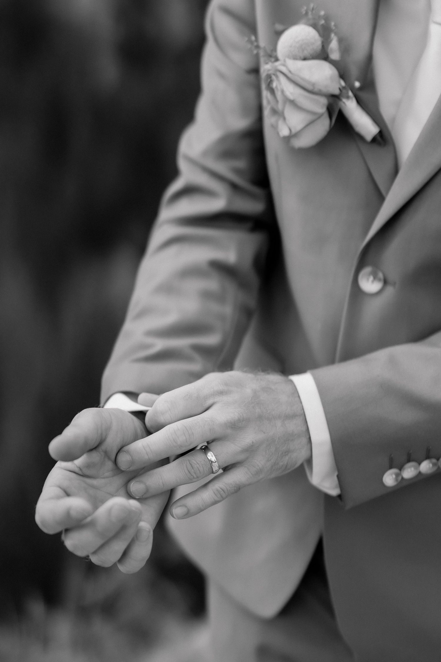 Black and white photo of groom placing wedding ring on bride's finger, wearing suit with boutonniere.