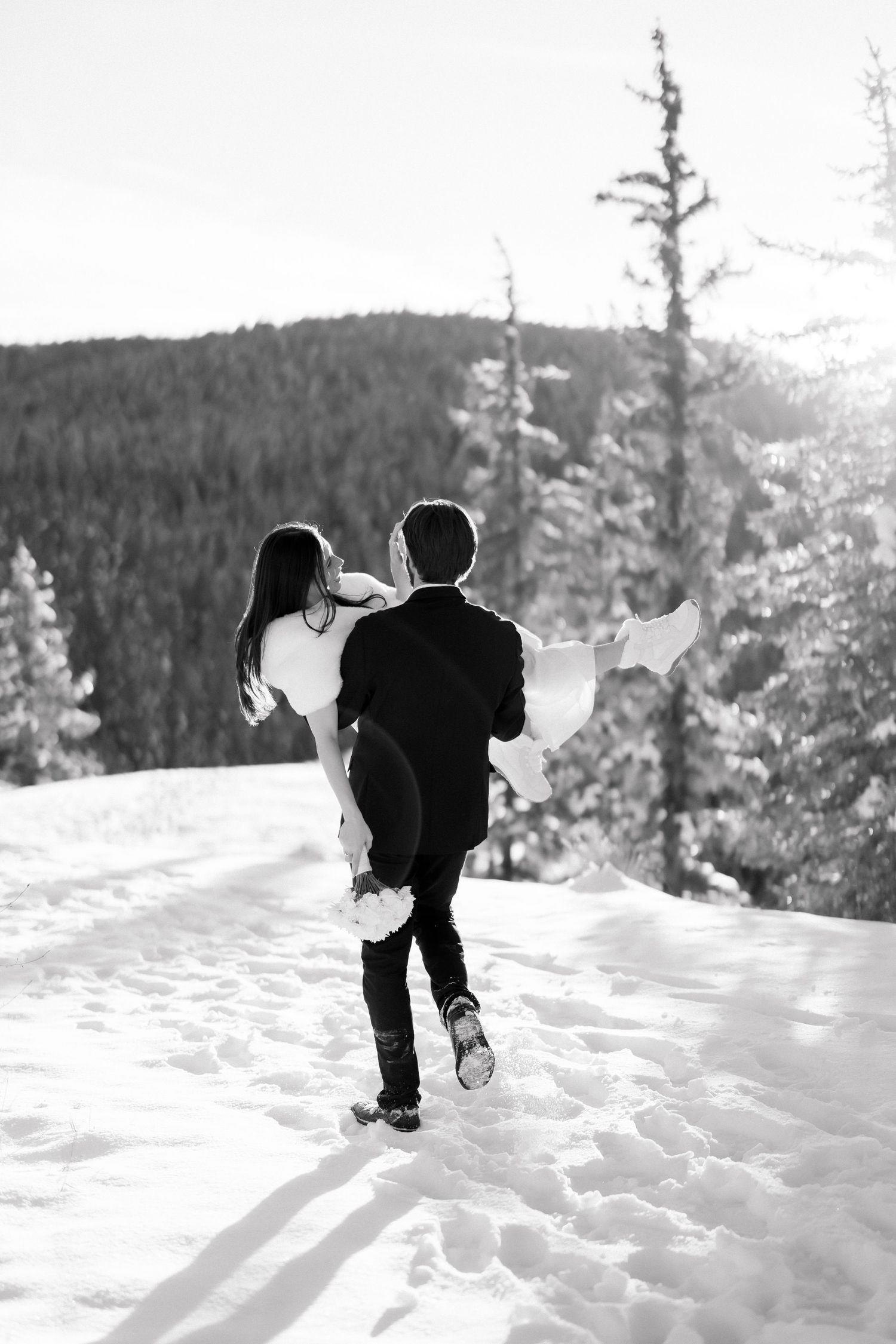 Black and white image of groom carrying bride through a snowy forest trail in winter wedding.