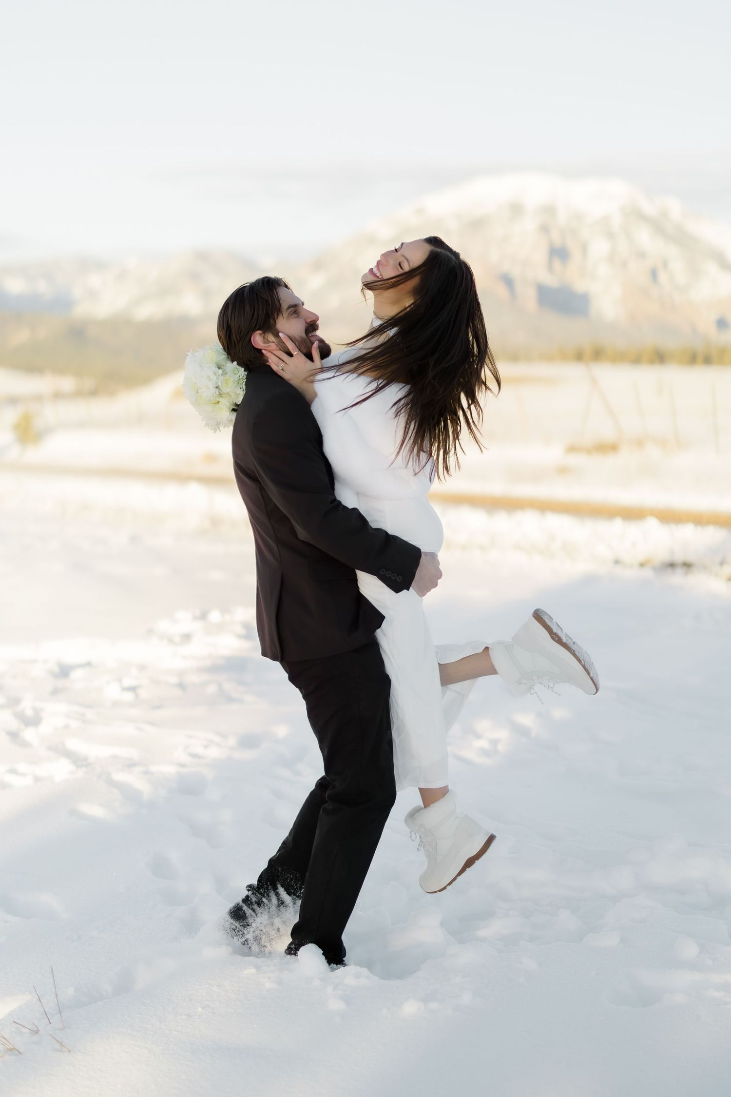 Groom lifts laughing bride in snowy mountain landscape during winter wedding.