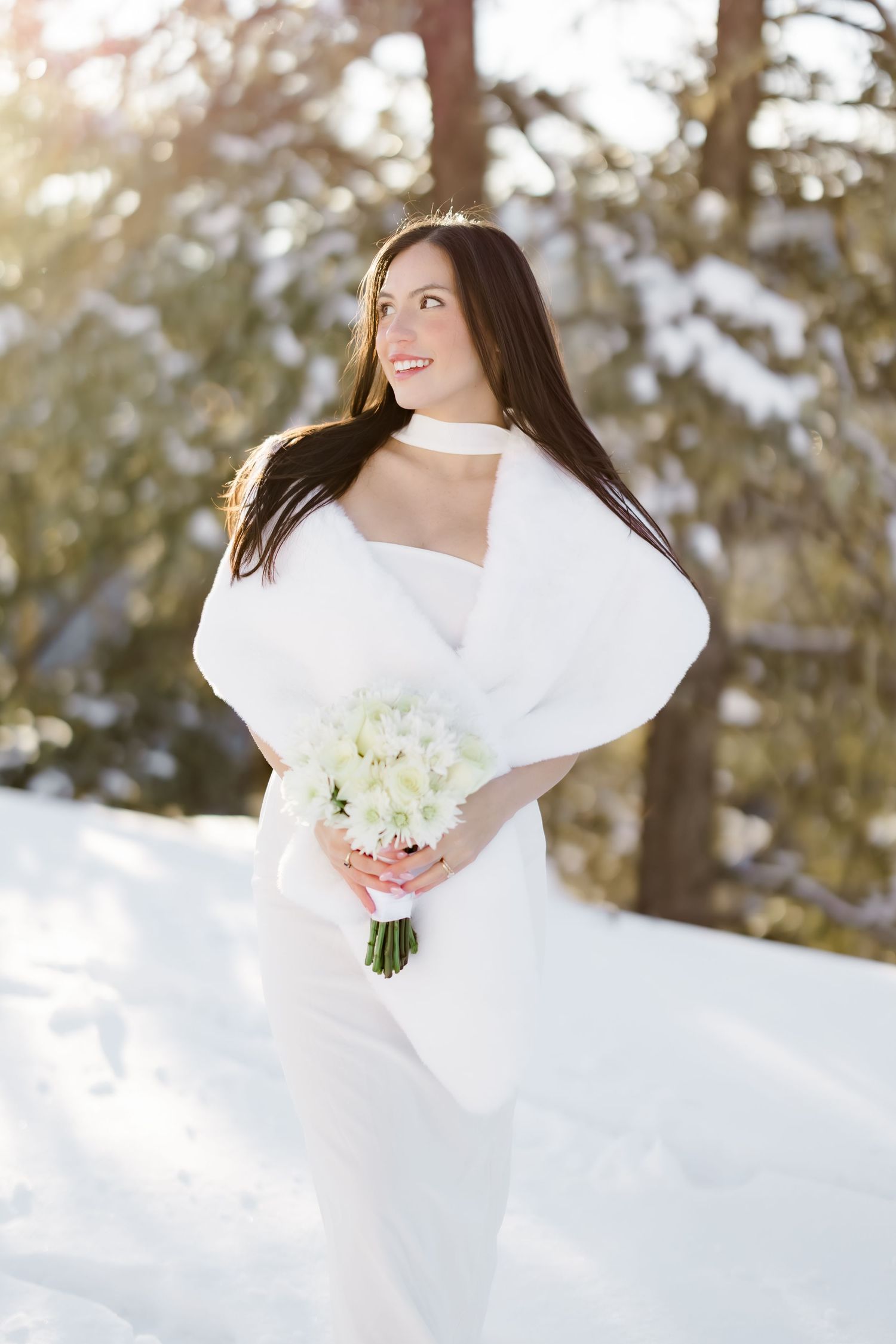 Smiling bride in white gown and fur wrap holding a bouquet in a snowy winter forest setting.