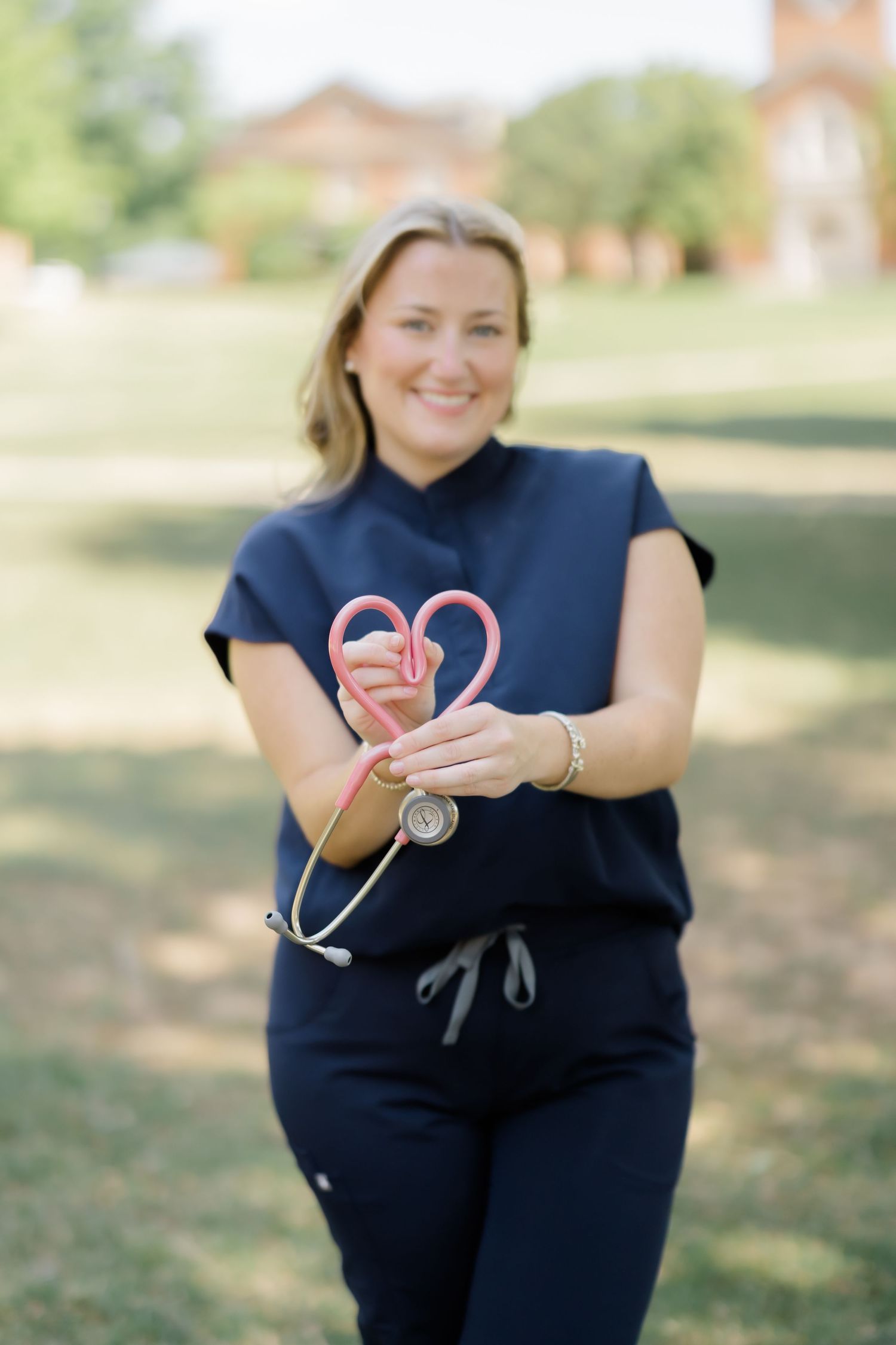 Smiling nurse in navy scrubs holds a pink stethoscope shaped into a heart outdoors on a sunny day.