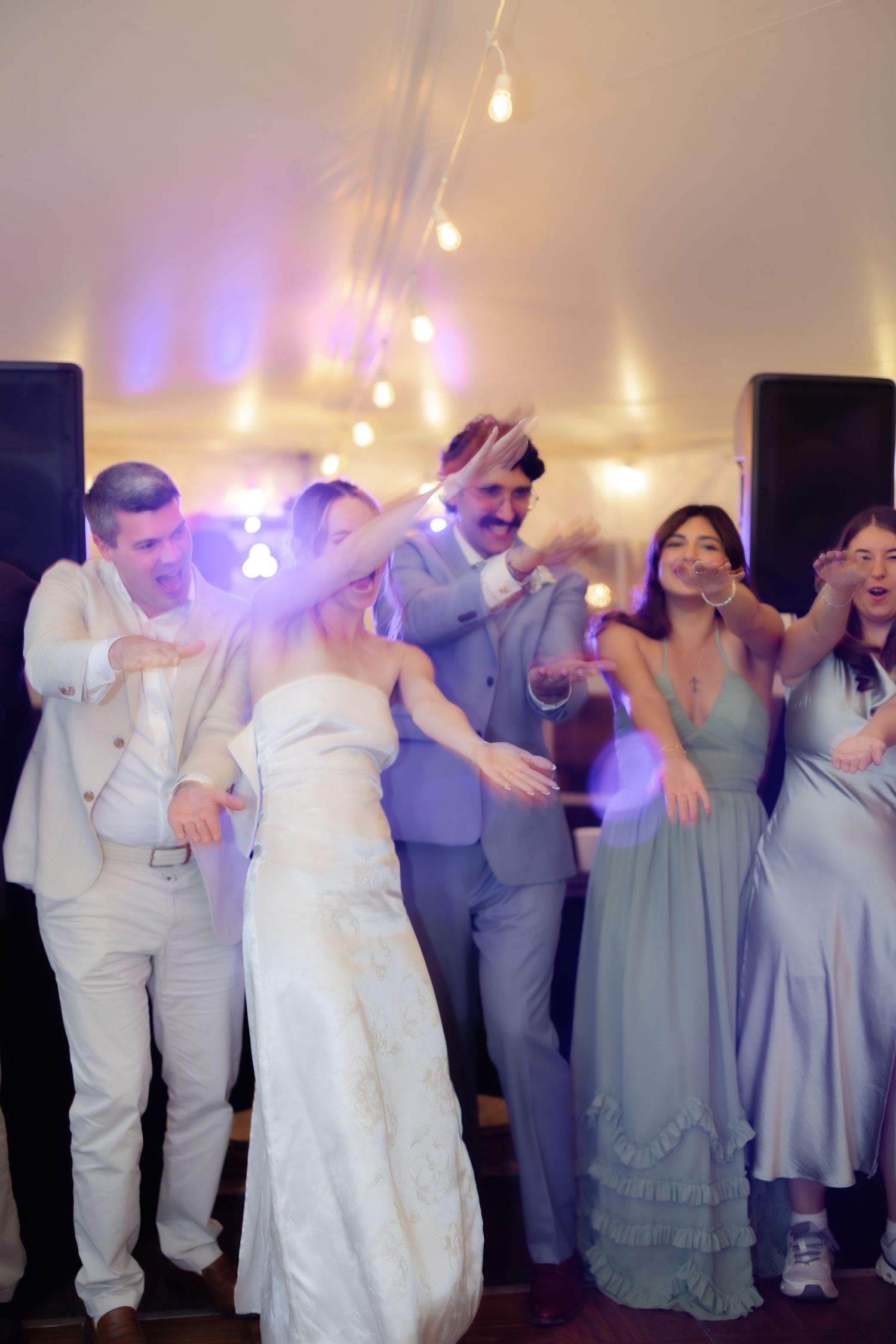 Bride and groom dancing joyfully with wedding guests at a reception venue with warm string lights.