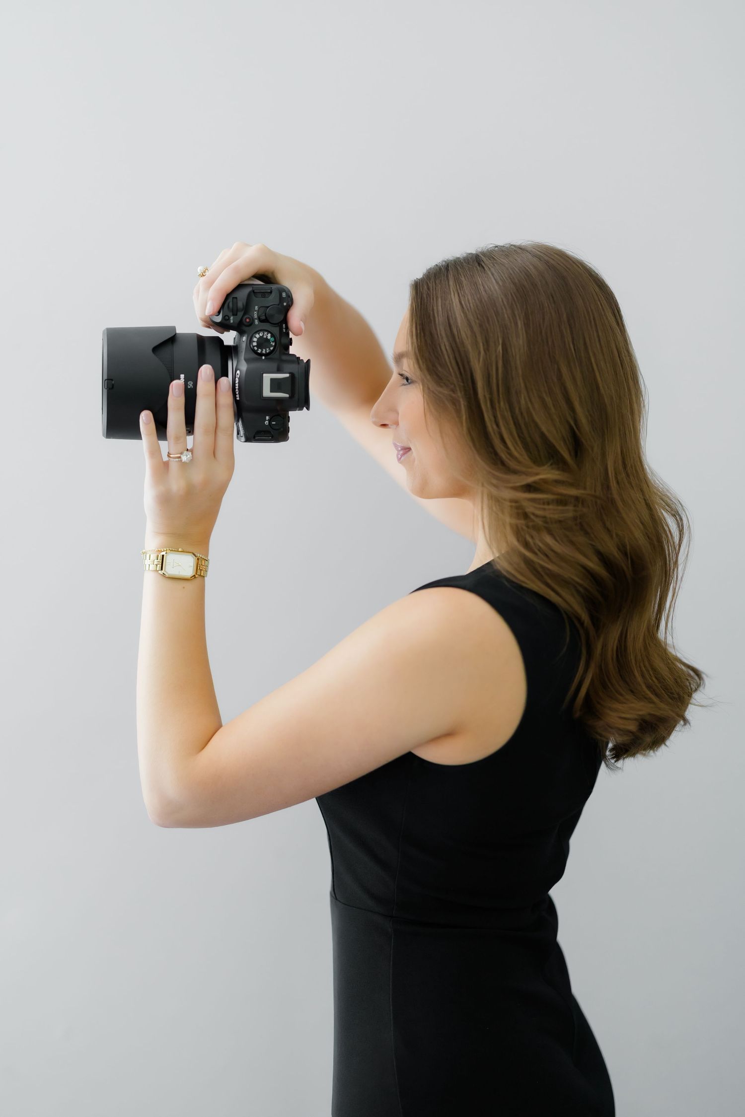 Woman in black dress photographing with a DSLR camera, wearing a gold watch and ring, side profile view.