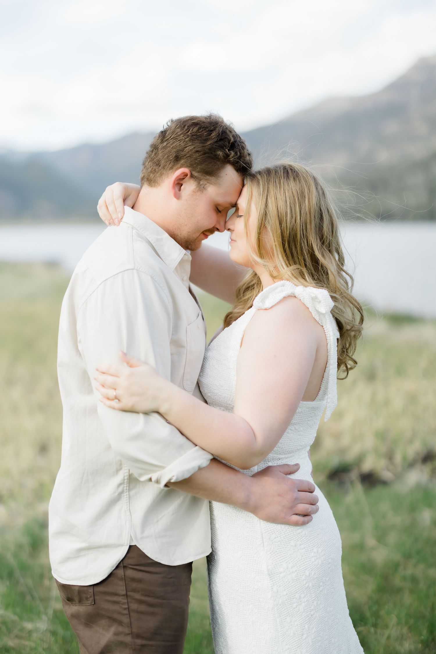 Couple embracing forehead to forehead in white dress and linen shirt with mountain lake backdrop.