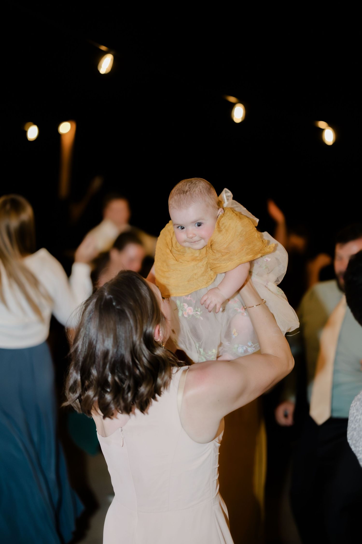 Woman in white dress lifts smiling baby in yellow outfit on a wedding reception dance floor.