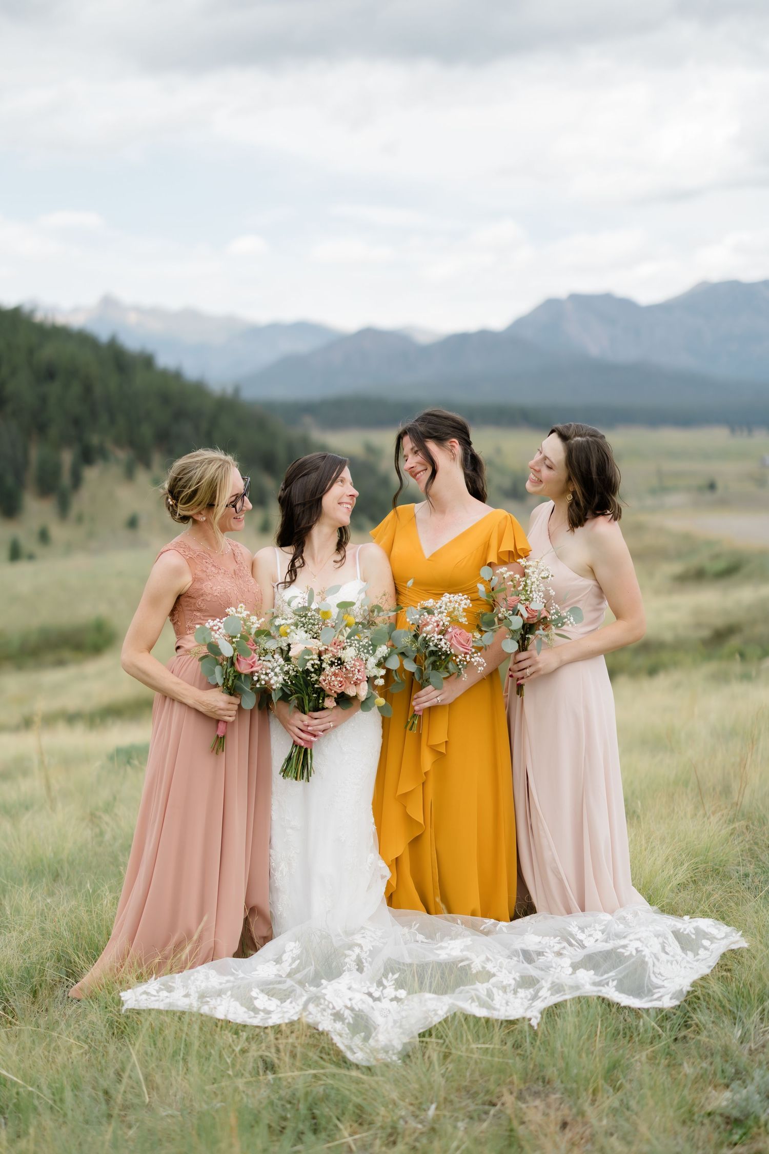 Bride and three bridesmaids in blush and mustard gowns laughing in a mountain meadow with bouquets.