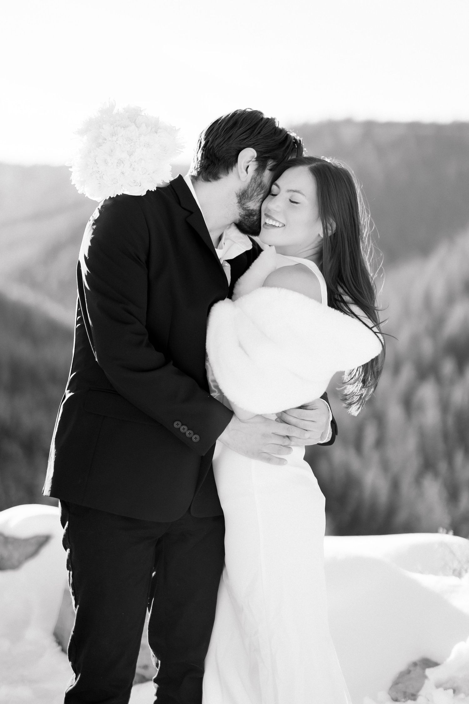 Black and white photo of groom kissing smiling bride on cheek in snowy mountain setting.