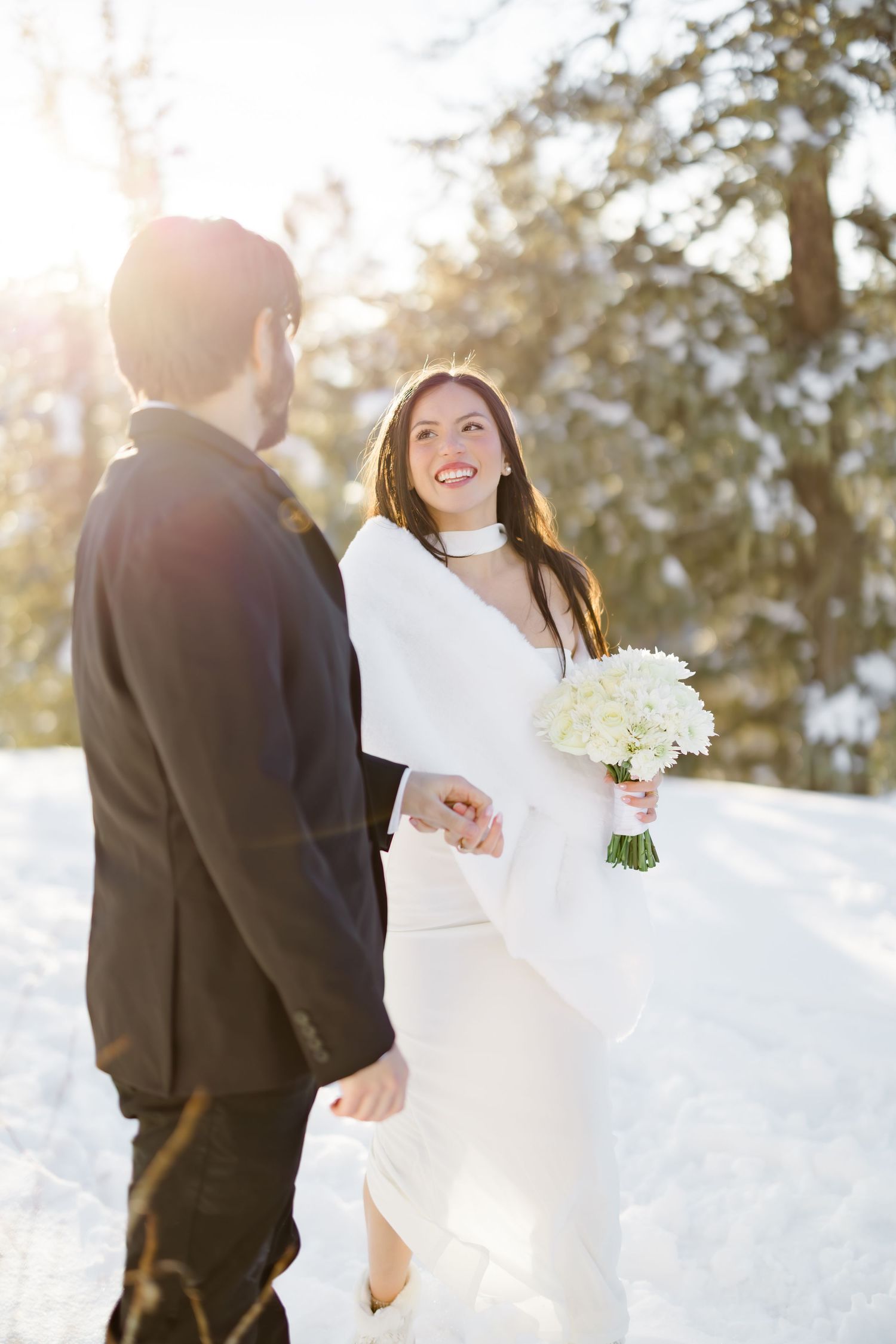Smiling bride in white dress holds bouquet while groom in black suit holds her hand in snow.