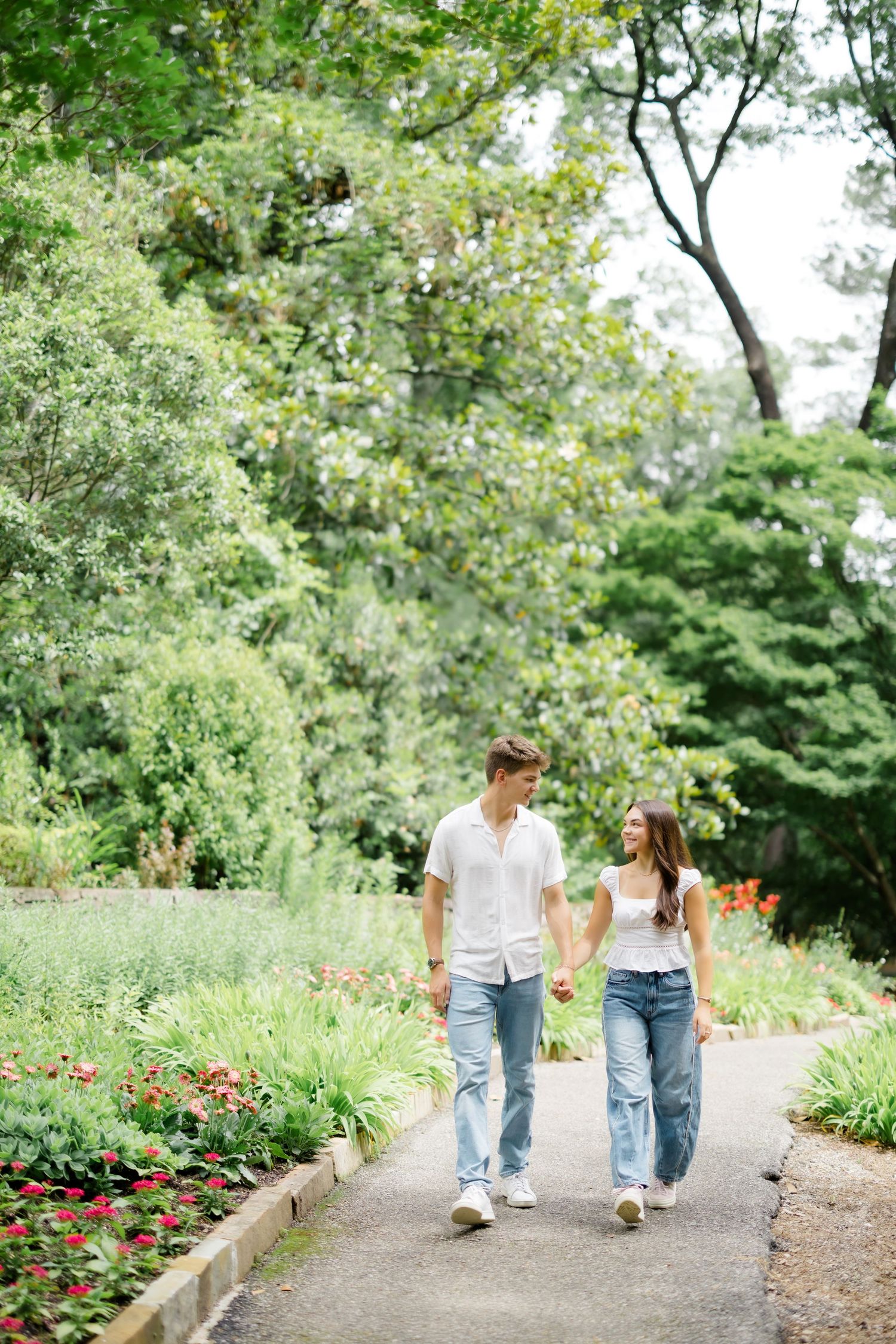 A happy couple holds hands while walking along a garden path surrounded by lush green trees and colorful flowers.