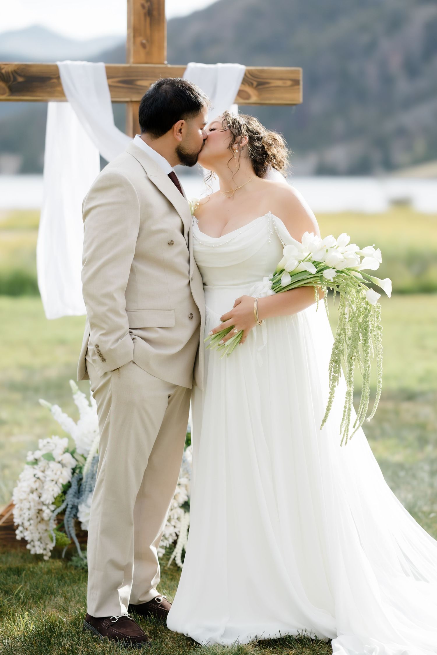 Bride and groom share a kiss before rustic wooden cross altar with mountain backdrop.