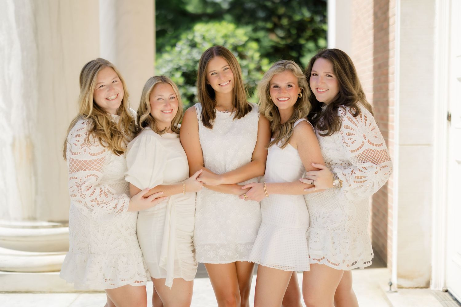 Five young women in white lace dresses pose together smiling between elegant columns outdoors.
