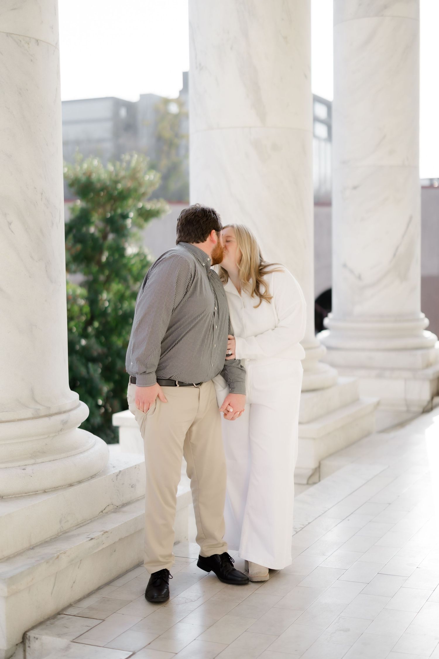 A couple shares a romantic kiss between elegant marble columns during an engagement photo session.