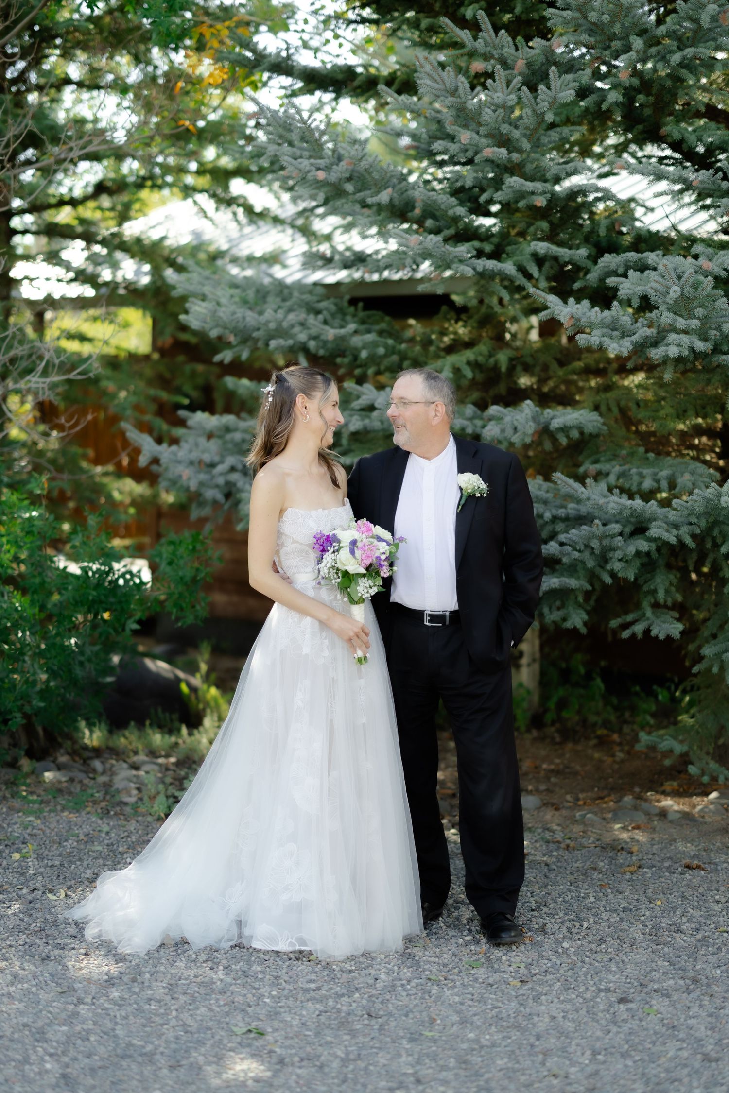 Bride in white gown holding bouquet stands with groom in black suit among evergreen trees.