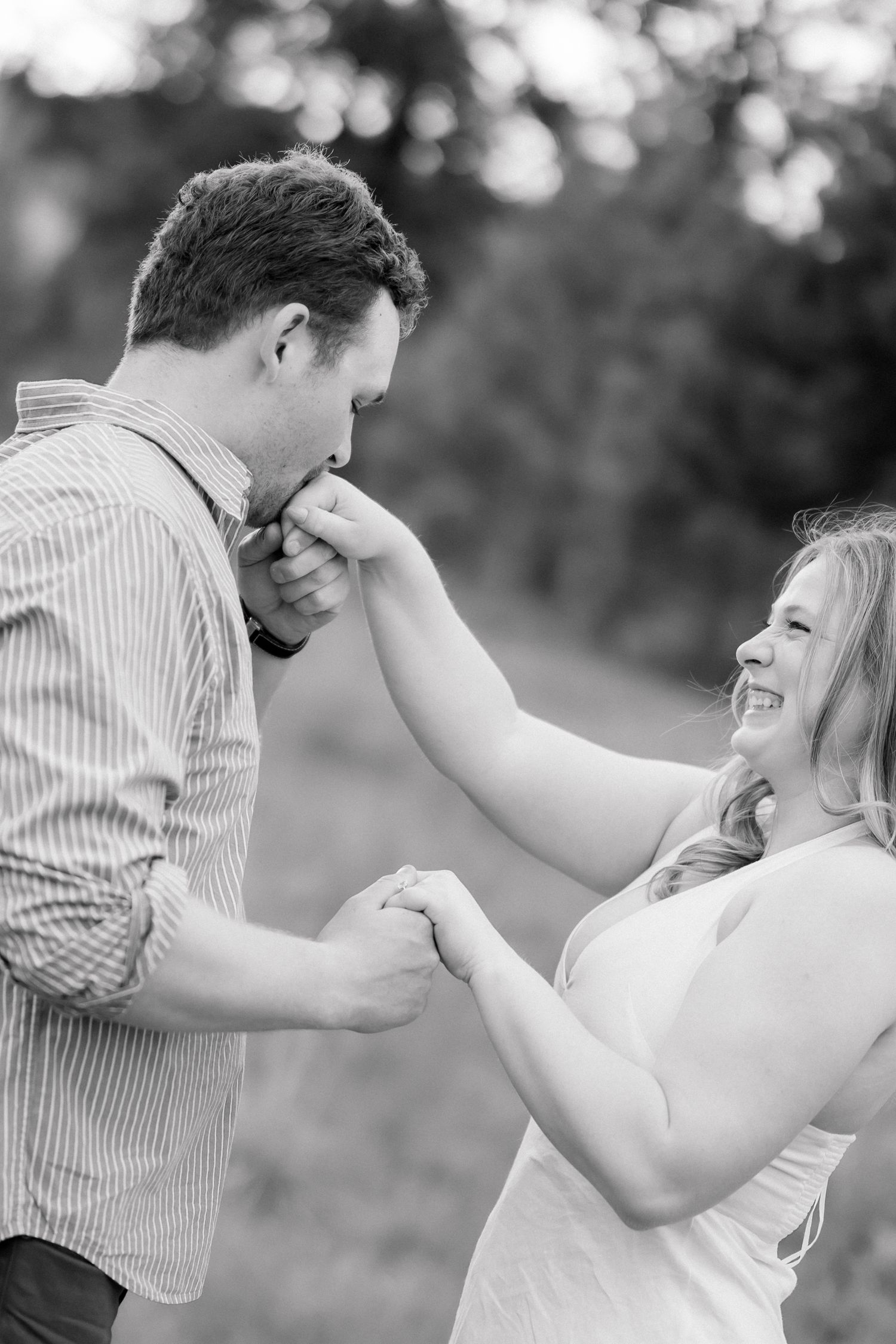 Man kisses woman's hand while she laughs joyfully in black and white engagement portrait.