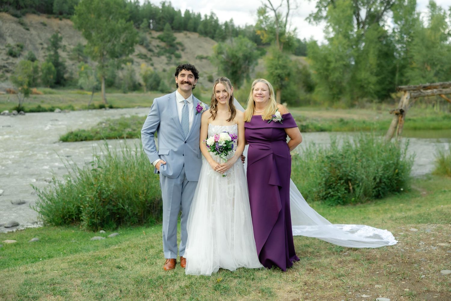 Bride in white gown poses with groom in gray suit and woman in purple dress by a river.