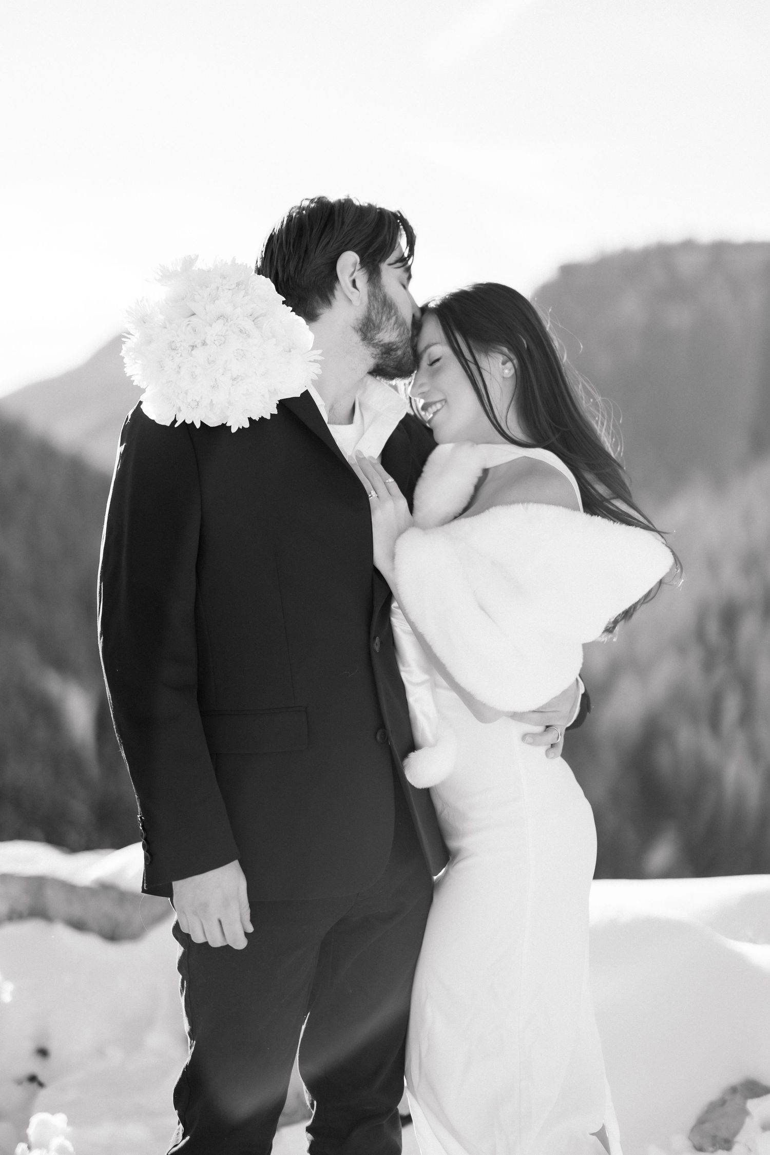 Black and white intimate portrait of newlyweds embracing closely against snowy mountain scenery.
