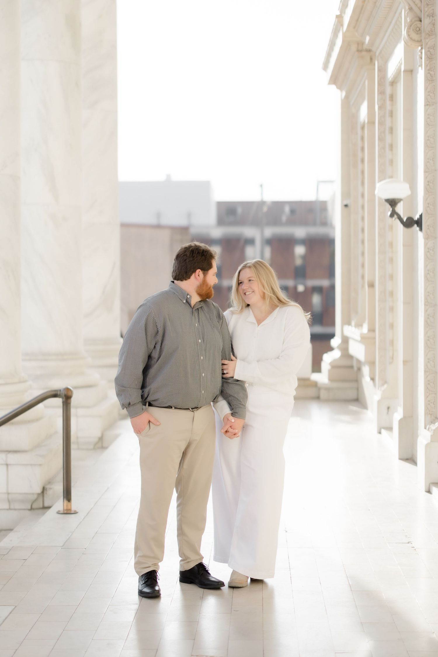 A smiling couple holds hands and gazes at each other between grand white columns.