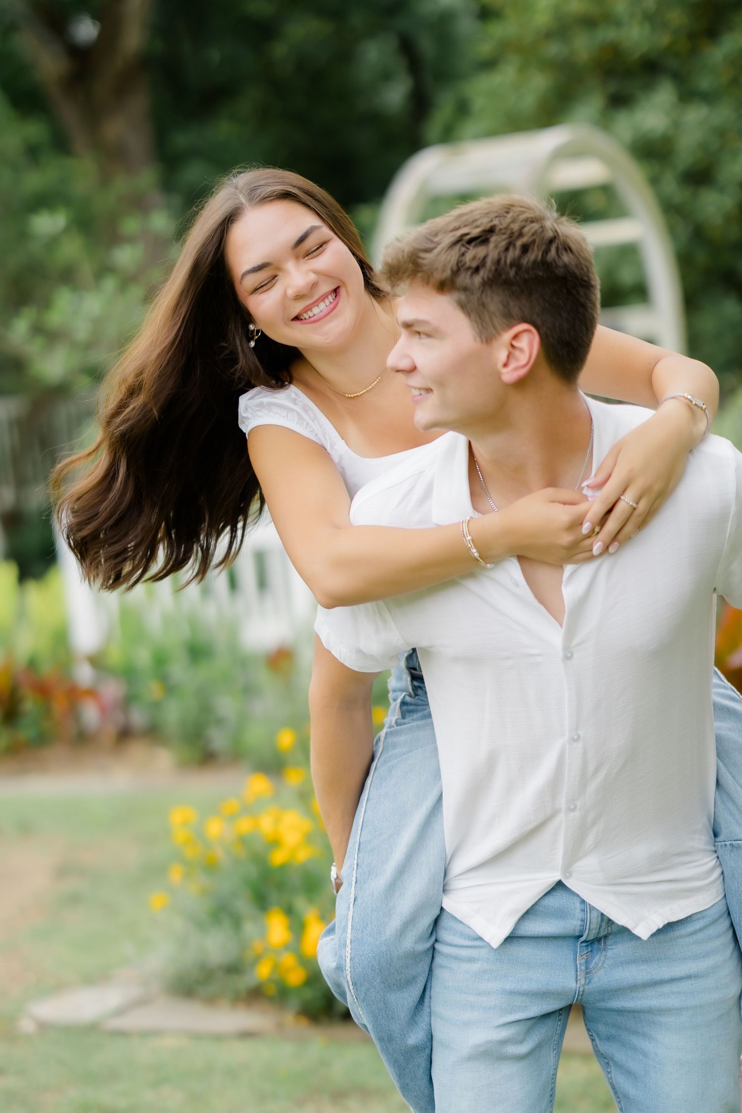 A happy young couple enjoys a piggyback ride outdoors, smiling in a lush green park with yellow flowers.